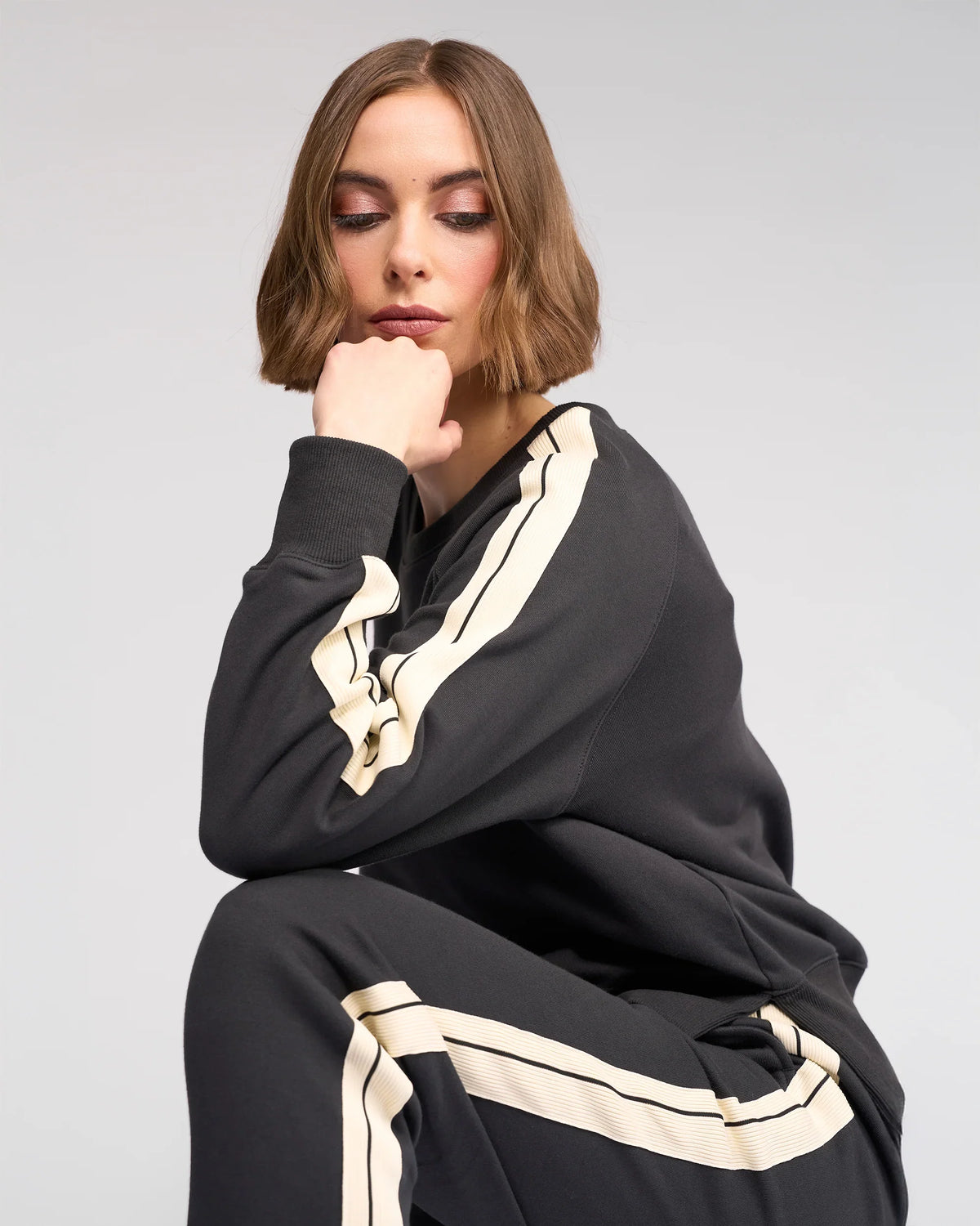 A woman with short brown hair sits in a thoughtful pose, resting her chin on her hand. She wears a dark tracksuit with light-colored stripes along the sleeves and pants. The background is plain and light-colored.