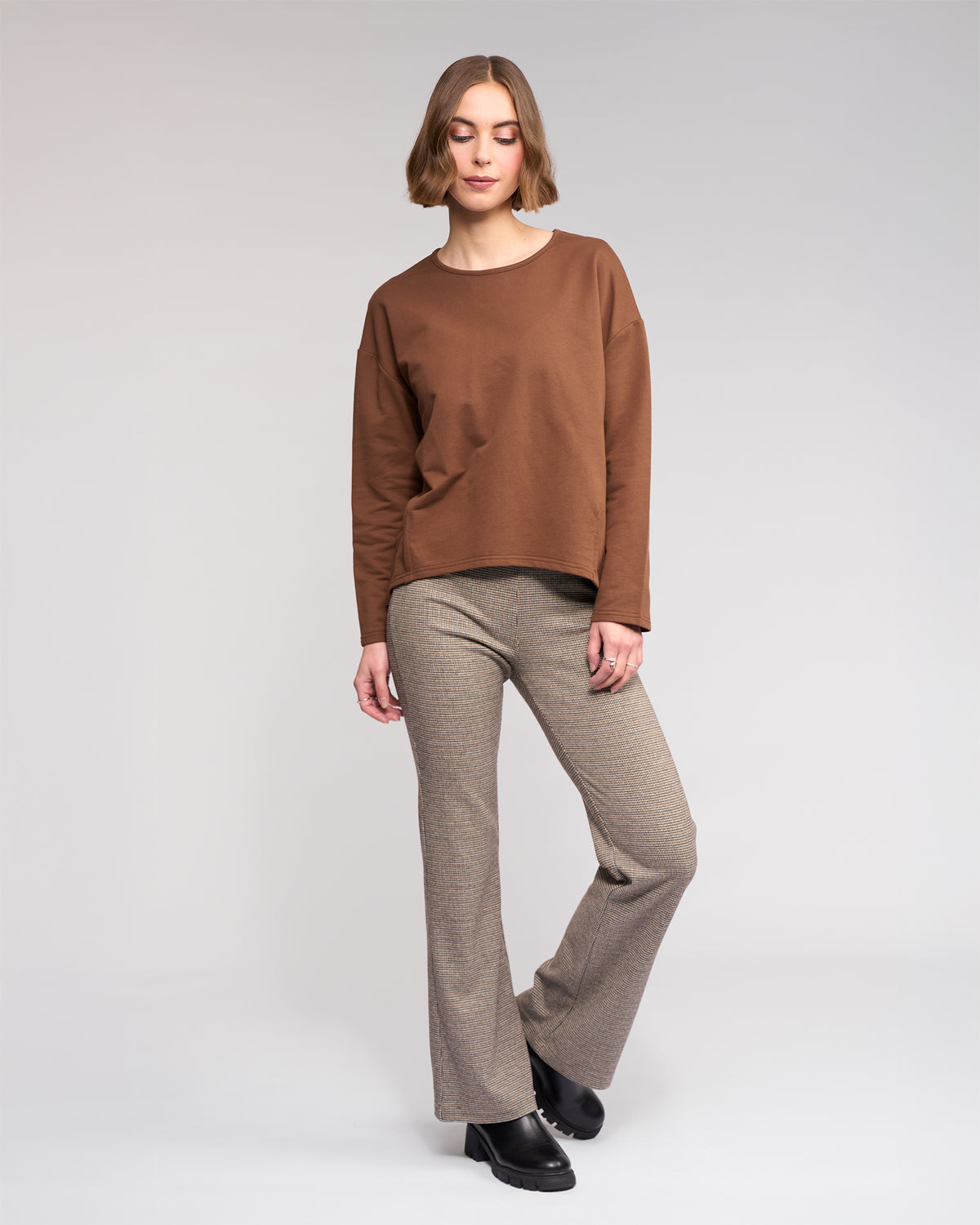 A woman with short light brown hair poses relaxedly against a plain background, wearing the Knewe Label Harley Top in Cedar, beige plaid flared pants, and black shoes. One leg is slightly bent.