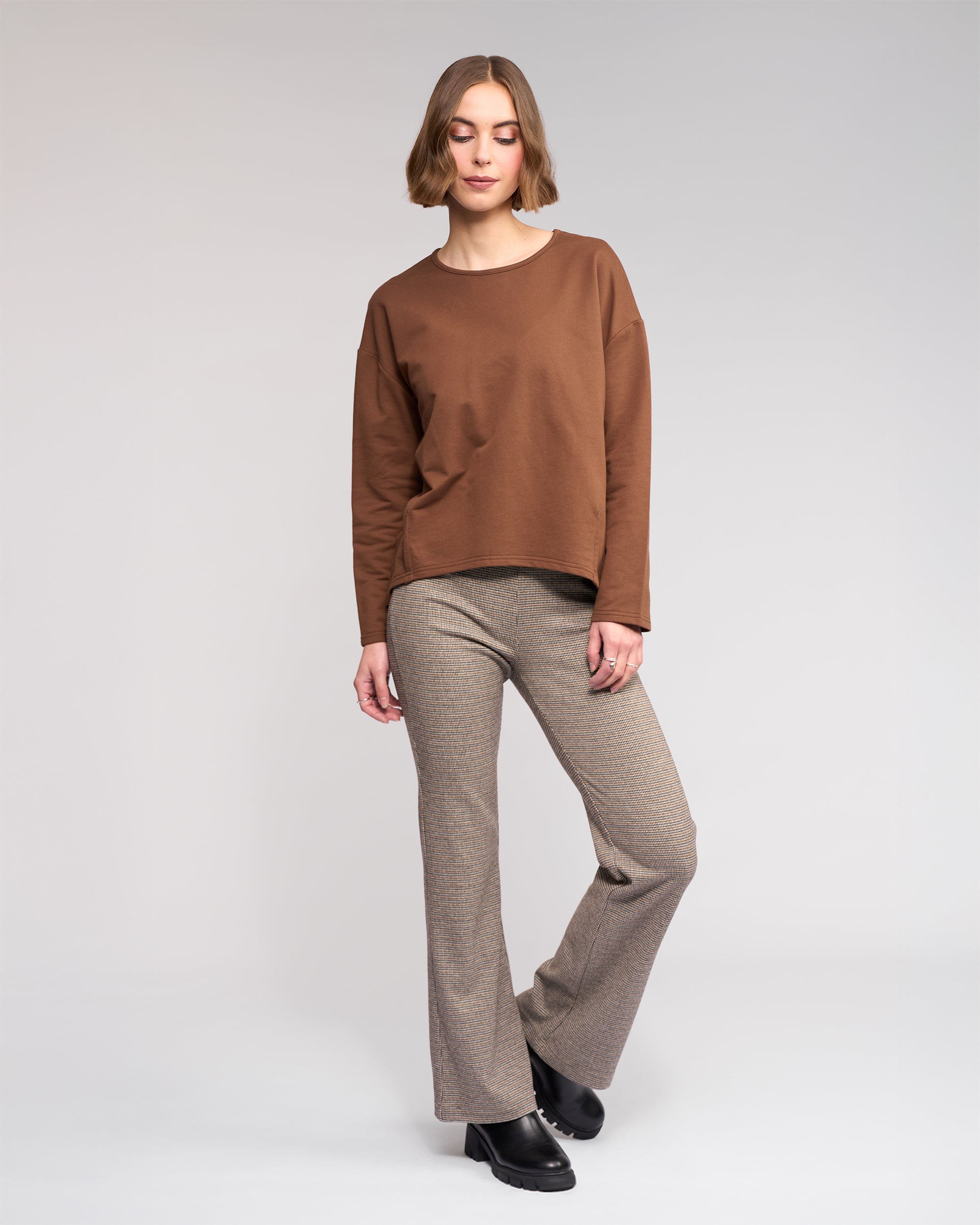 A woman with short light brown hair poses relaxedly against a plain background, wearing the Knewe Label Harley Top in Cedar, beige plaid flared pants, and black shoes. One leg is slightly bent.