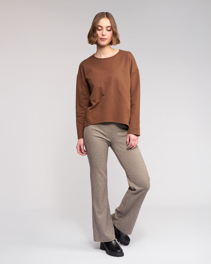 A woman with short light brown hair poses relaxedly against a plain background, wearing the Knewe Label Harley Top in Cedar, beige plaid flared pants, and black shoes. One leg is slightly bent.