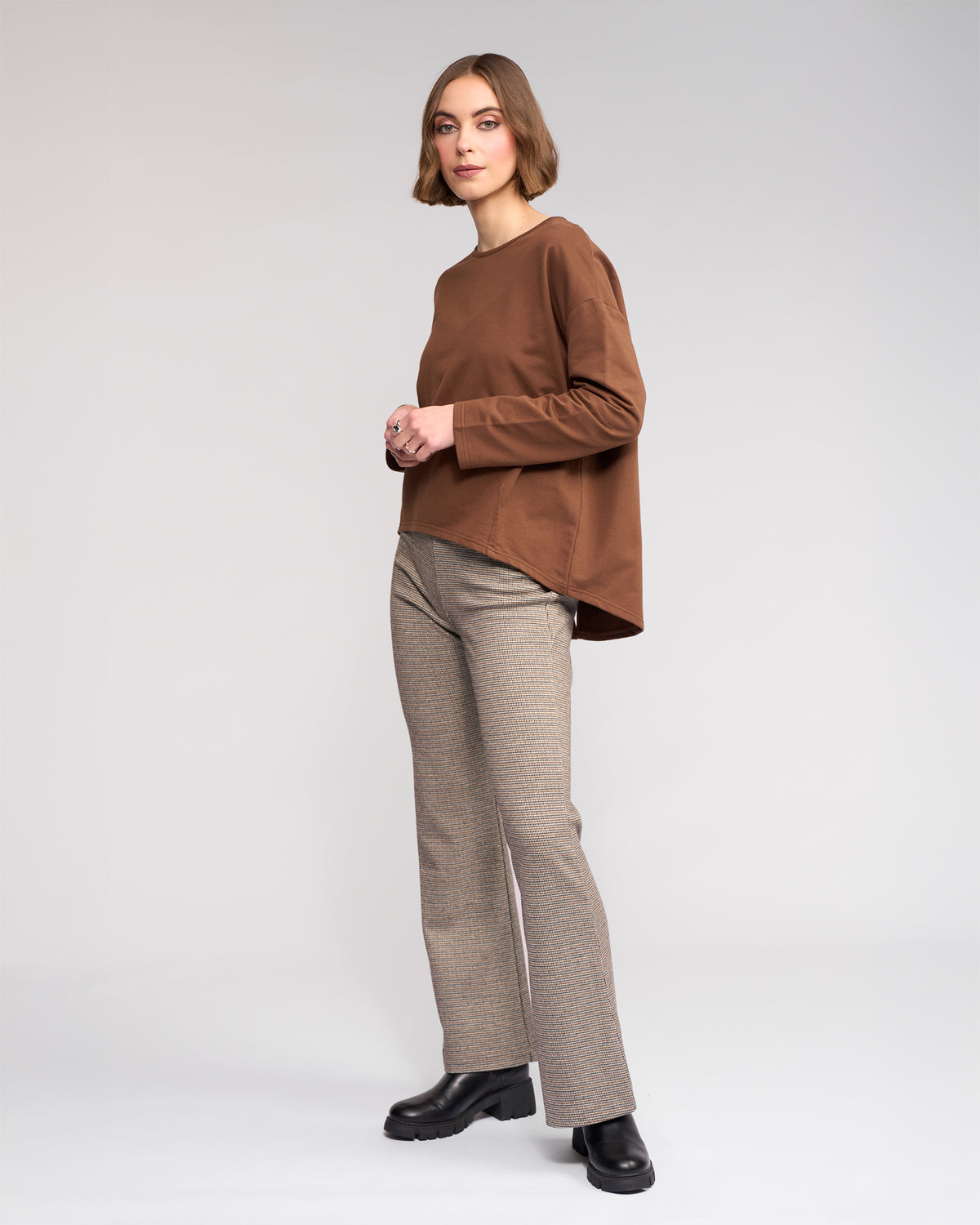 A woman with short brown hair stands against a light gray background, wearing the Knewe Label Harley Top in Cedar, beige plaid pants, and black platform shoes. She faces the camera with a neutral expression.