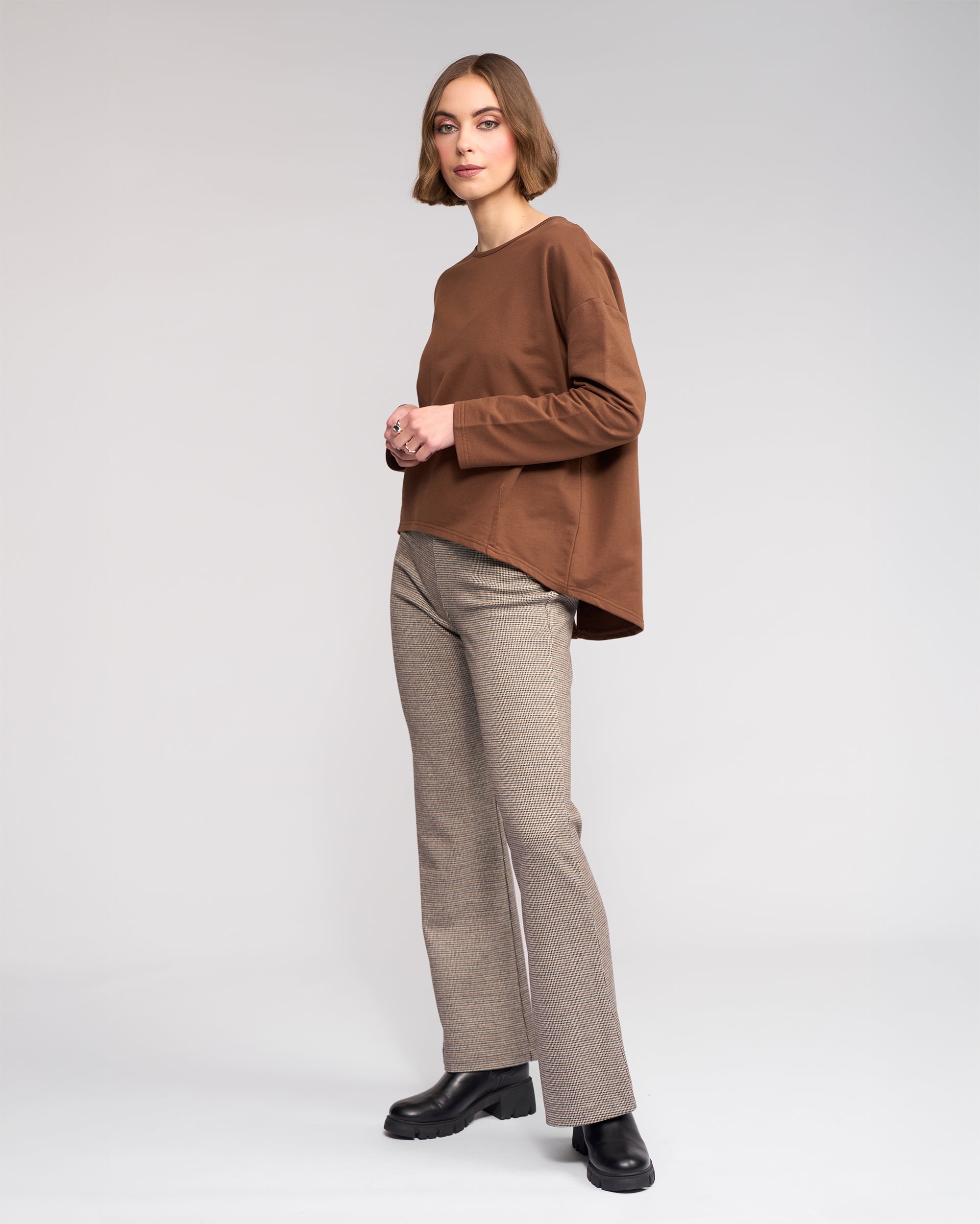 A woman with short brown hair stands against a light gray background, wearing the Knewe Label Harley Top in Cedar, beige plaid pants, and black platform shoes. She faces the camera with a neutral expression.