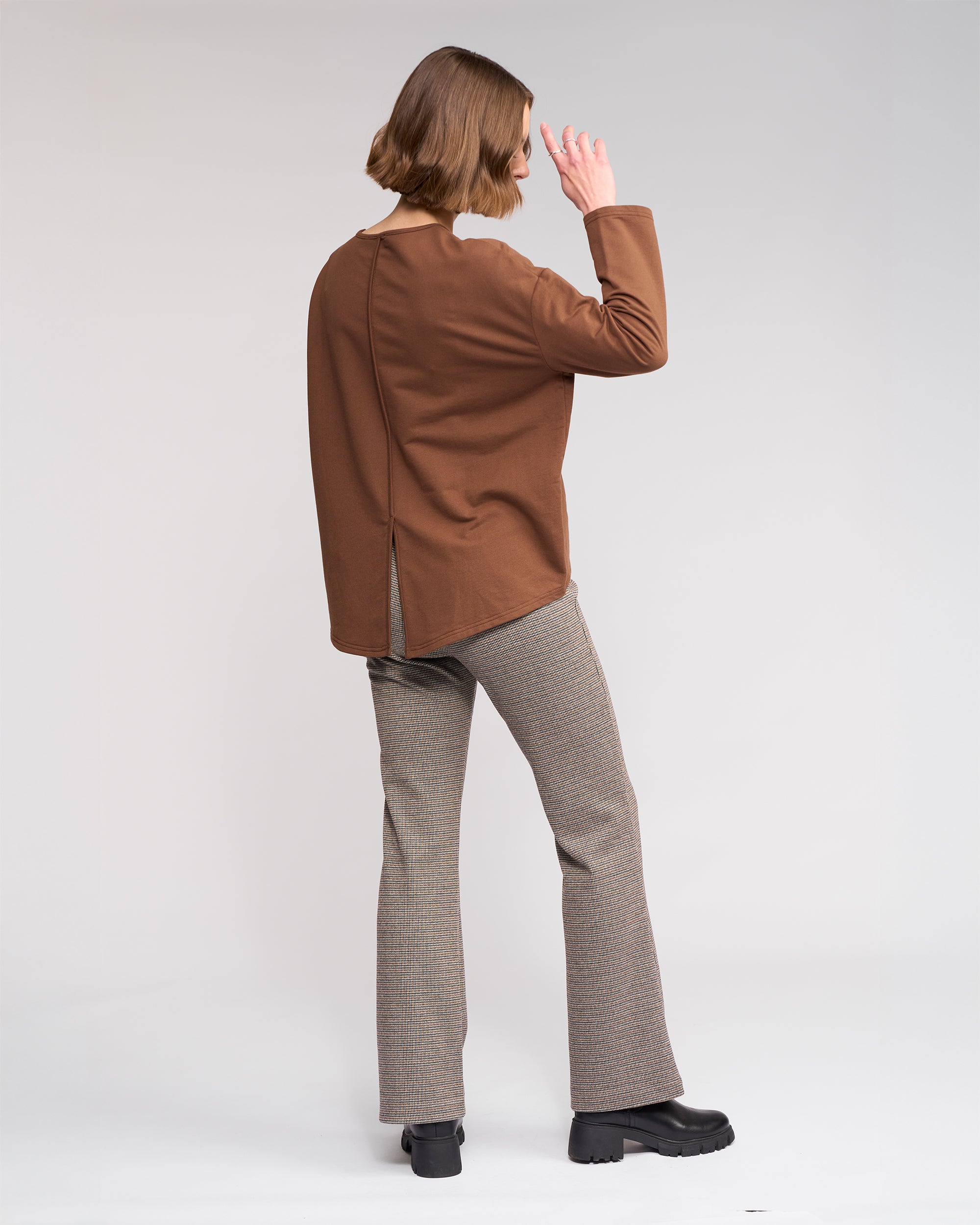 A person with short hair faces away, wearing Knewe Label's Harley Top in Cedar—a golden brown oversized knit with a back slit—and gray plaid pants, raising one hand near their head against a white background.