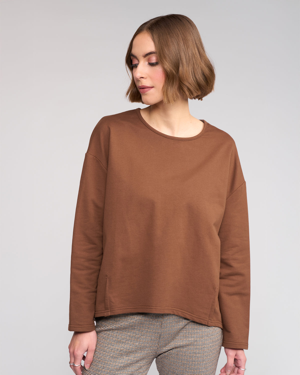 A woman with short, light brown hair wears the Knewe Label Harley Top in Cedar and plaid pants against a neutral background, looking downward with a relaxed expression.