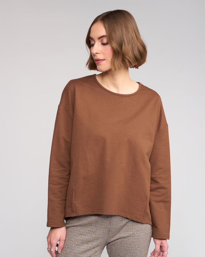 A woman with short, light brown hair wears the Knewe Label Harley Top in Cedar and plaid pants against a neutral background, looking downward with a relaxed expression.