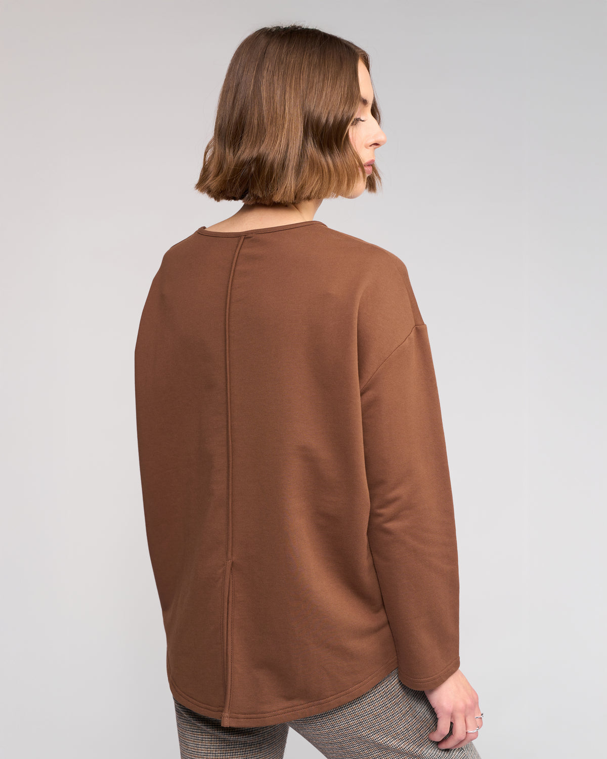 A woman with a short brown bob is shown from the back, wearing the Knewe Label Harley Top in Cedar and checked pants against a plain light background.