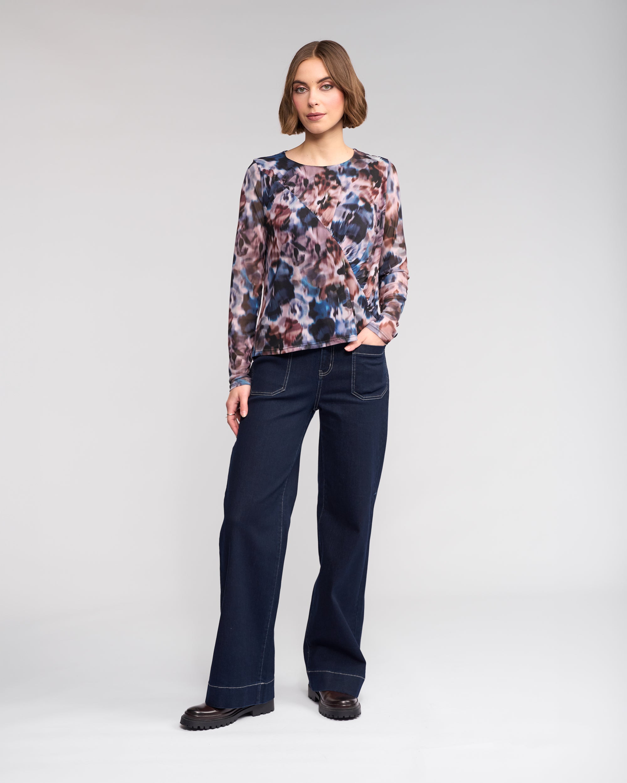 A woman stands against a plain background, wearing the Knewe Label Apex Top - Celine, long sleeve with an abstract print mesh, paired with dark wide-leg jeans and black shoes. One hand is in her pocket as she looks at the camera.