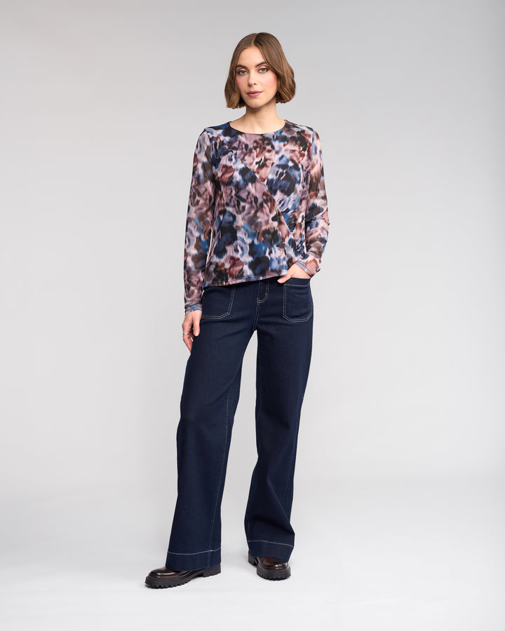 A woman stands against a plain background, wearing the Knewe Label Apex Top - Celine, long sleeve with an abstract print mesh, paired with dark wide-leg jeans and black shoes. One hand is in her pocket as she looks at the camera.