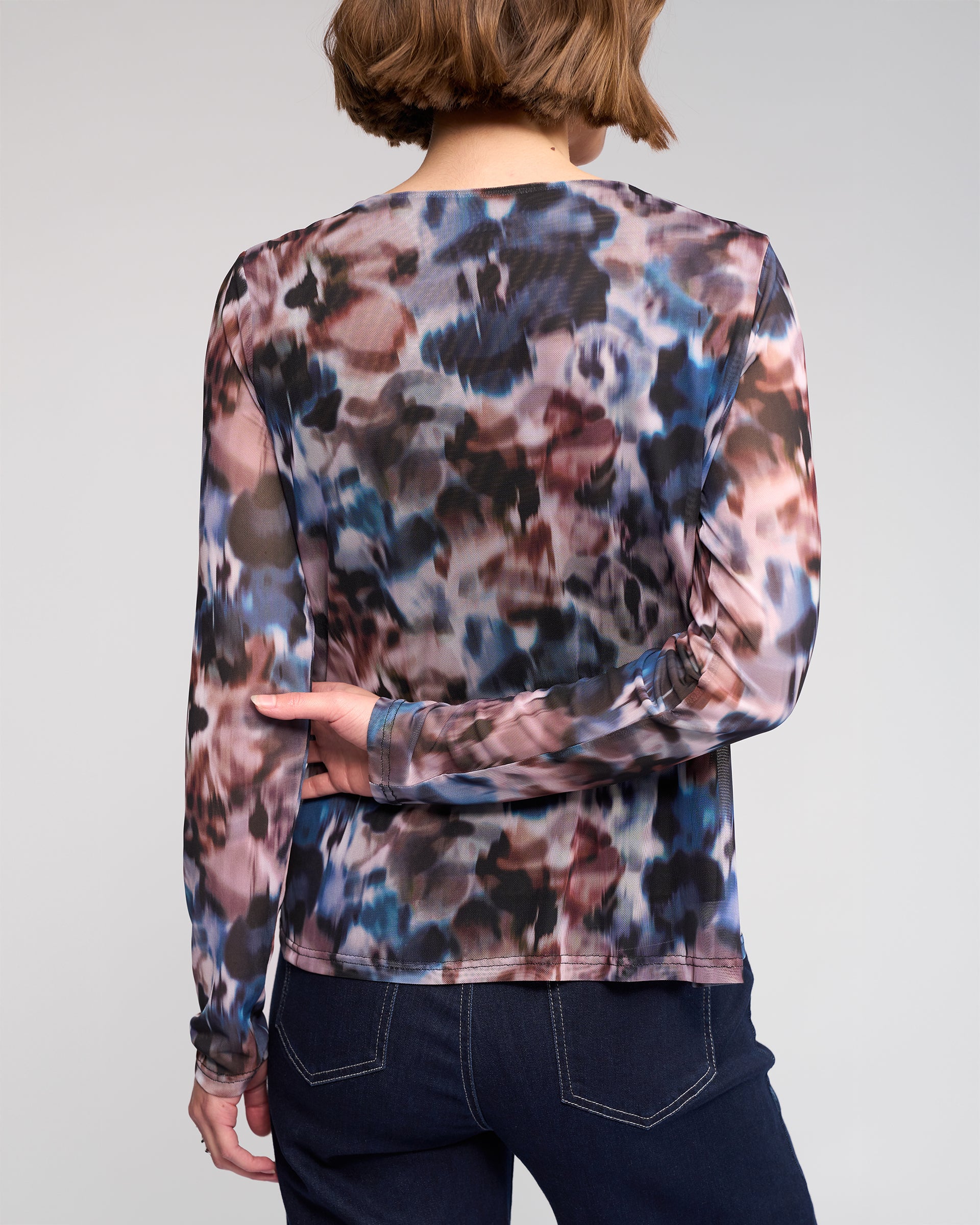 A person with short brown hair, seen from the back, wears the Knewe Label Apex Top - Celine—an abstract print mesh long sleeve—paired with dark blue jeans against a plain background.
