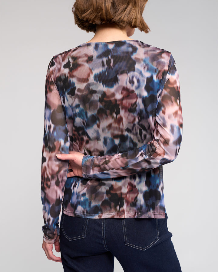 A person with short brown hair, seen from the back, wears the Knewe Label Apex Top - Celine—an abstract print mesh long sleeve—paired with dark blue jeans against a plain background.