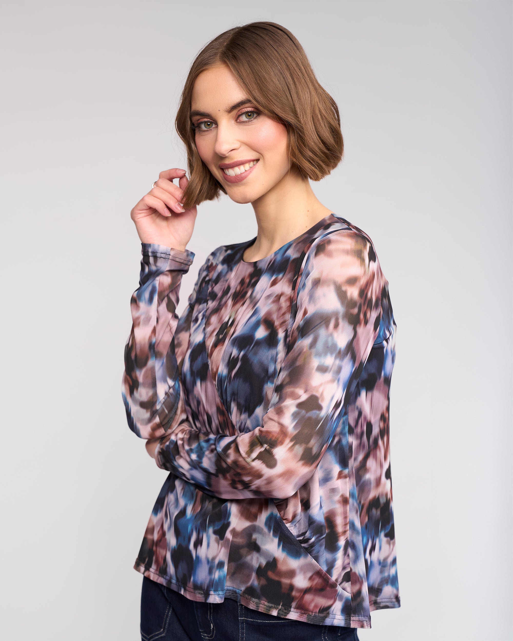 A woman with short brown hair smiles with one hand near her face, wearing the Apex Top - Celine by Knewe Label—an abstract print mesh long sleeve—paired with dark jeans, against a light gray background.