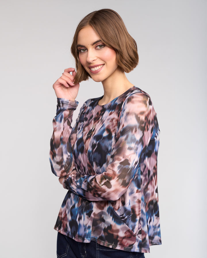 A woman with short brown hair smiles with one hand near her face, wearing the Apex Top - Celine by Knewe Label—an abstract print mesh long sleeve—paired with dark jeans, against a light gray background.