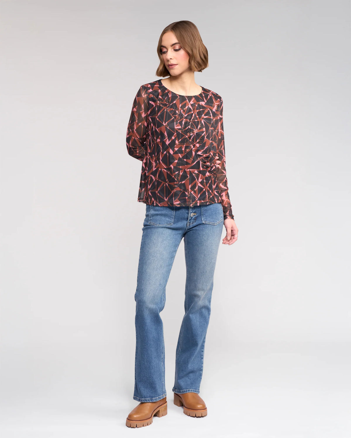 A woman with short, wavy hair looks down to her left, wearing the Knewe Label Apex Top - Dusk, a long sleeve mesh top in dark tones, paired with light blue jeans and brown platform shoes against a plain background.