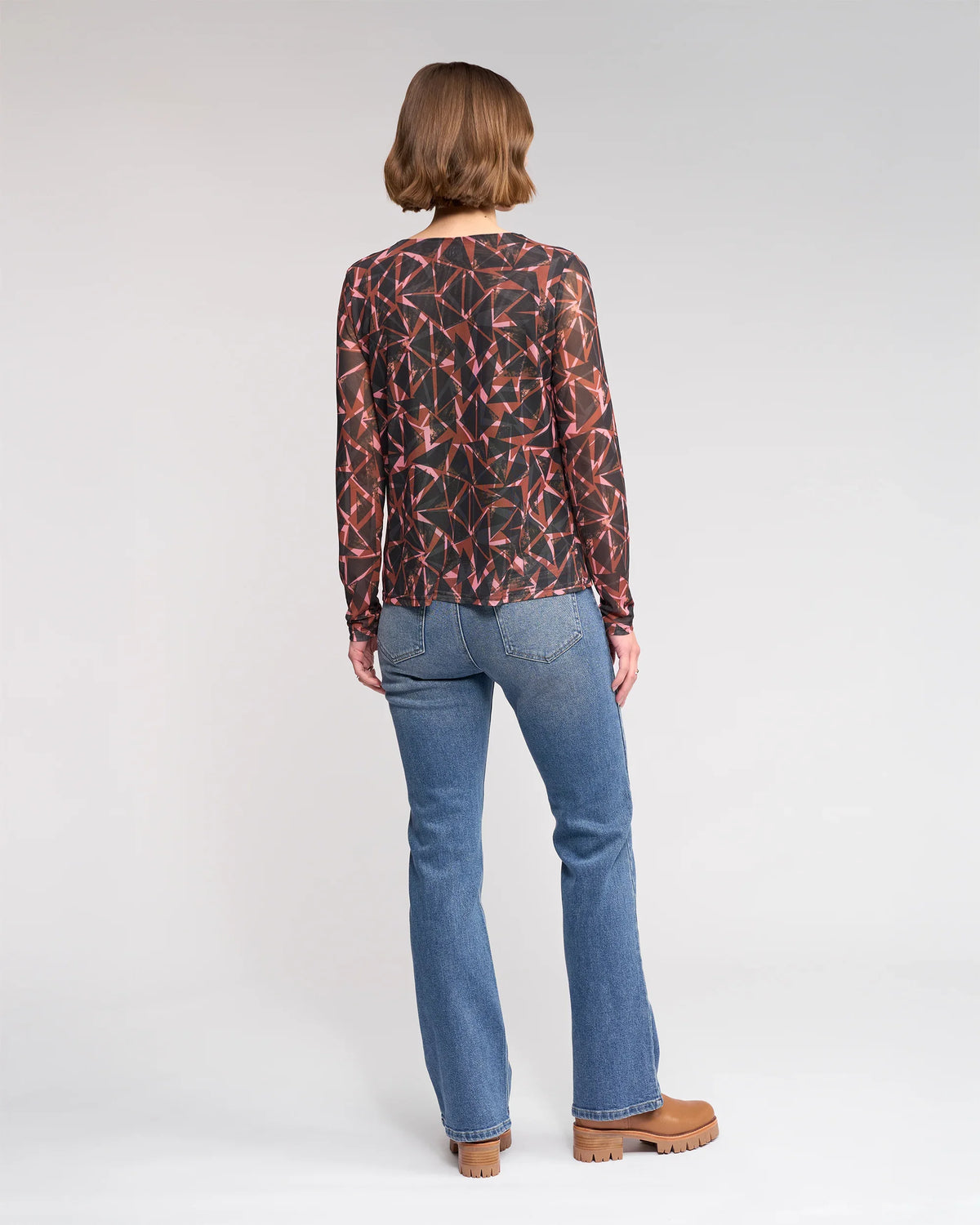 A person with short brown hair stands with their back to the camera, wearing the Knewe Label Apex Top - Dusk, blue jeans, and brown platform shoes against a plain light background.
