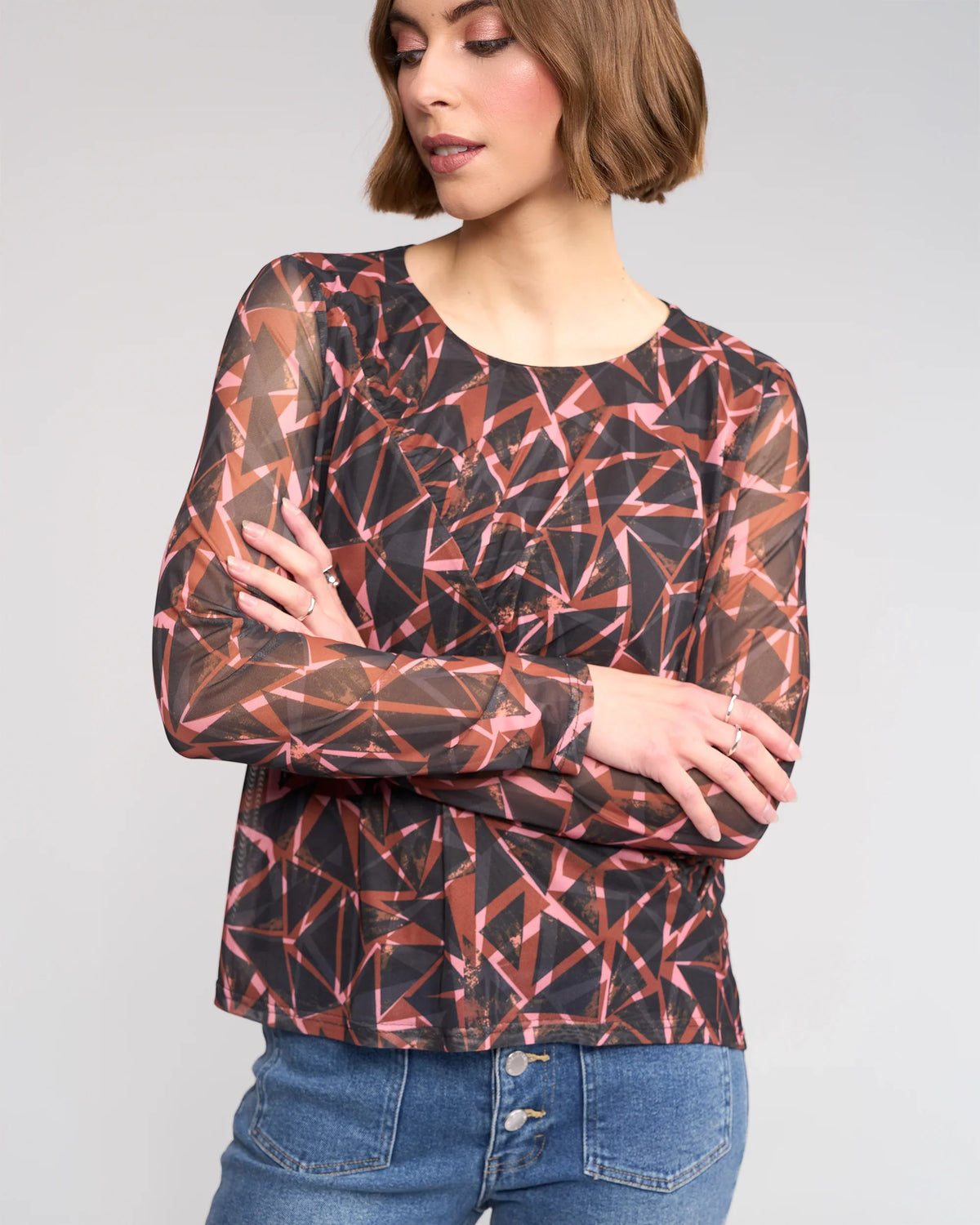A woman with short brown hair wears the Knewe Label Apex Top - Dusk, a long sleeve mesh top with red, pink, and black geometric dusk print, paired with high-waisted blue jeans. She stands with arms crossed, looking to the side.