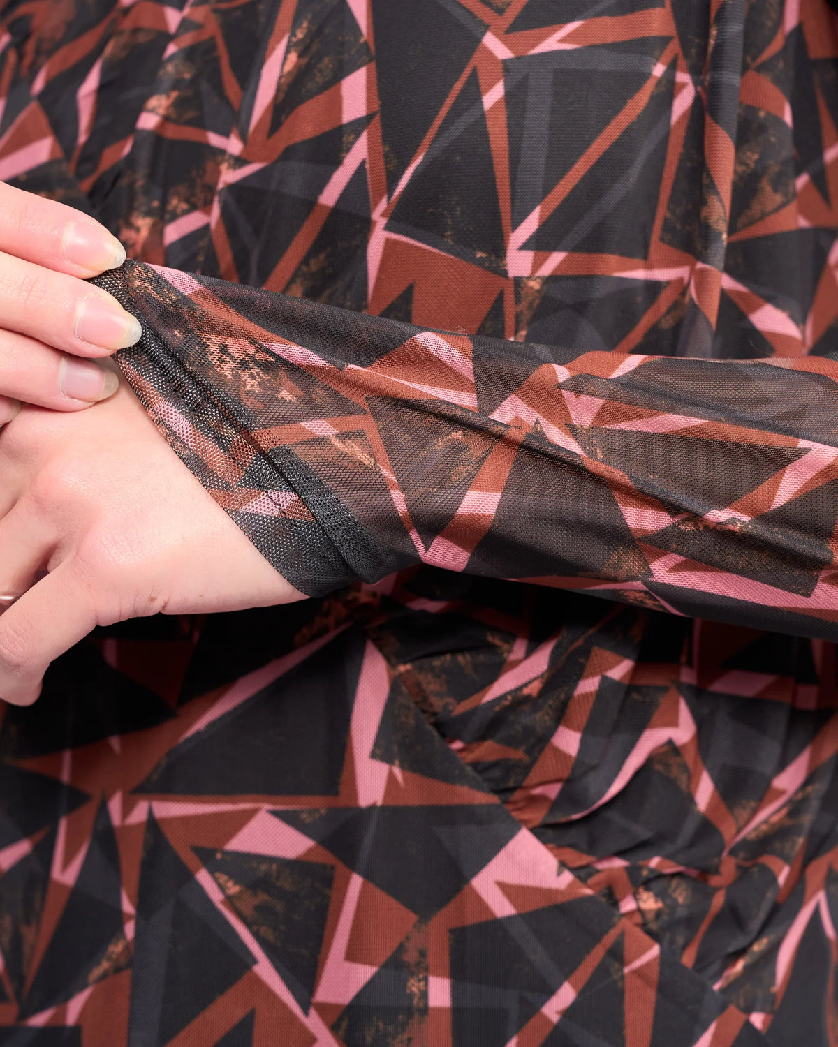 A close-up of a hand gently pulling the sleeve of Knewe Label's Apex Top - Dusk, a long sleeve mesh top in black, brown, and pink geometric print with soft, textured fabric.