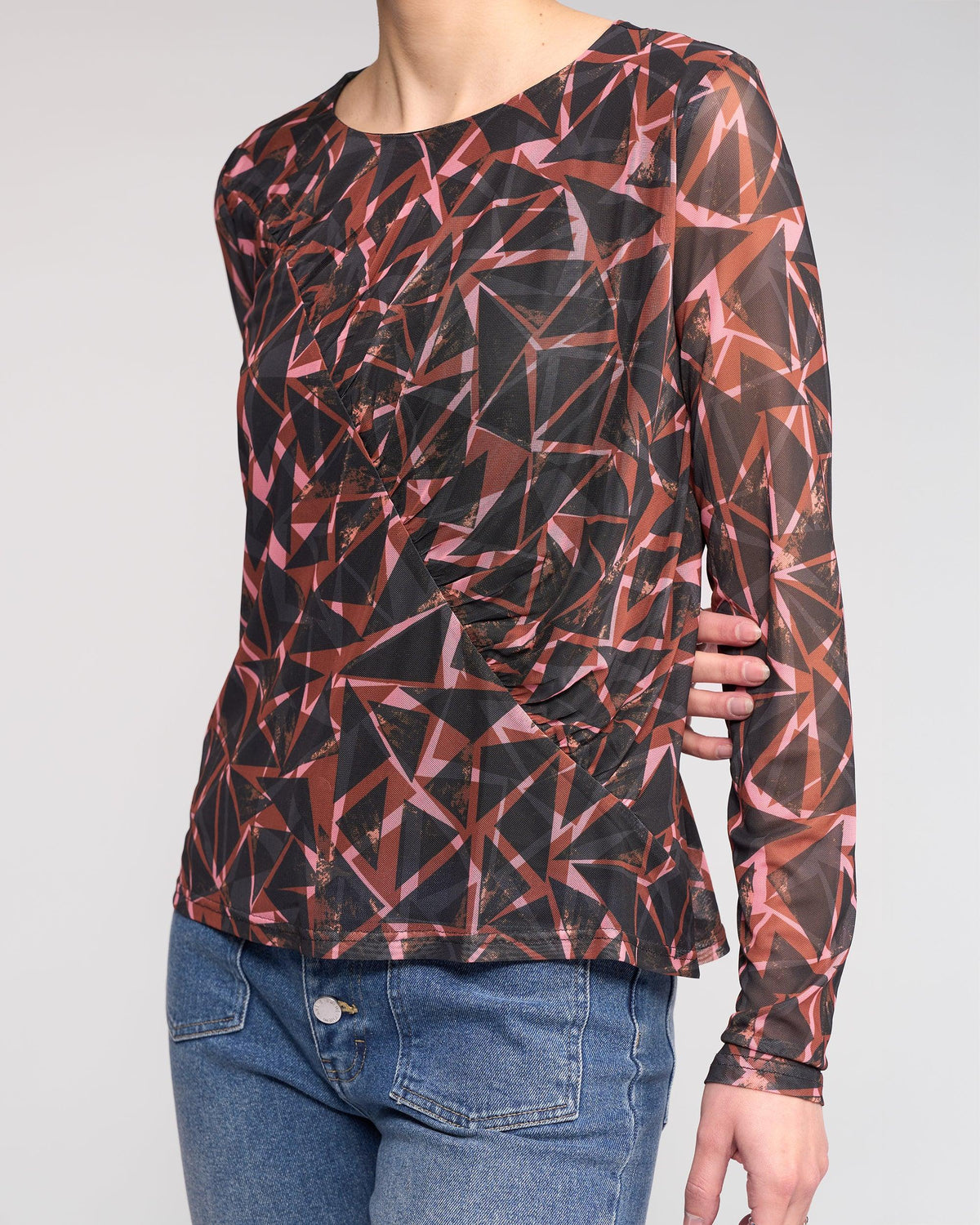 A person wears the Knewe Label Apex Top - Dusk, a long sleeve mesh top in a geometric red, pink, and black print, paired with blue jeans. Their head is out of frame and one hand rests on their hip.