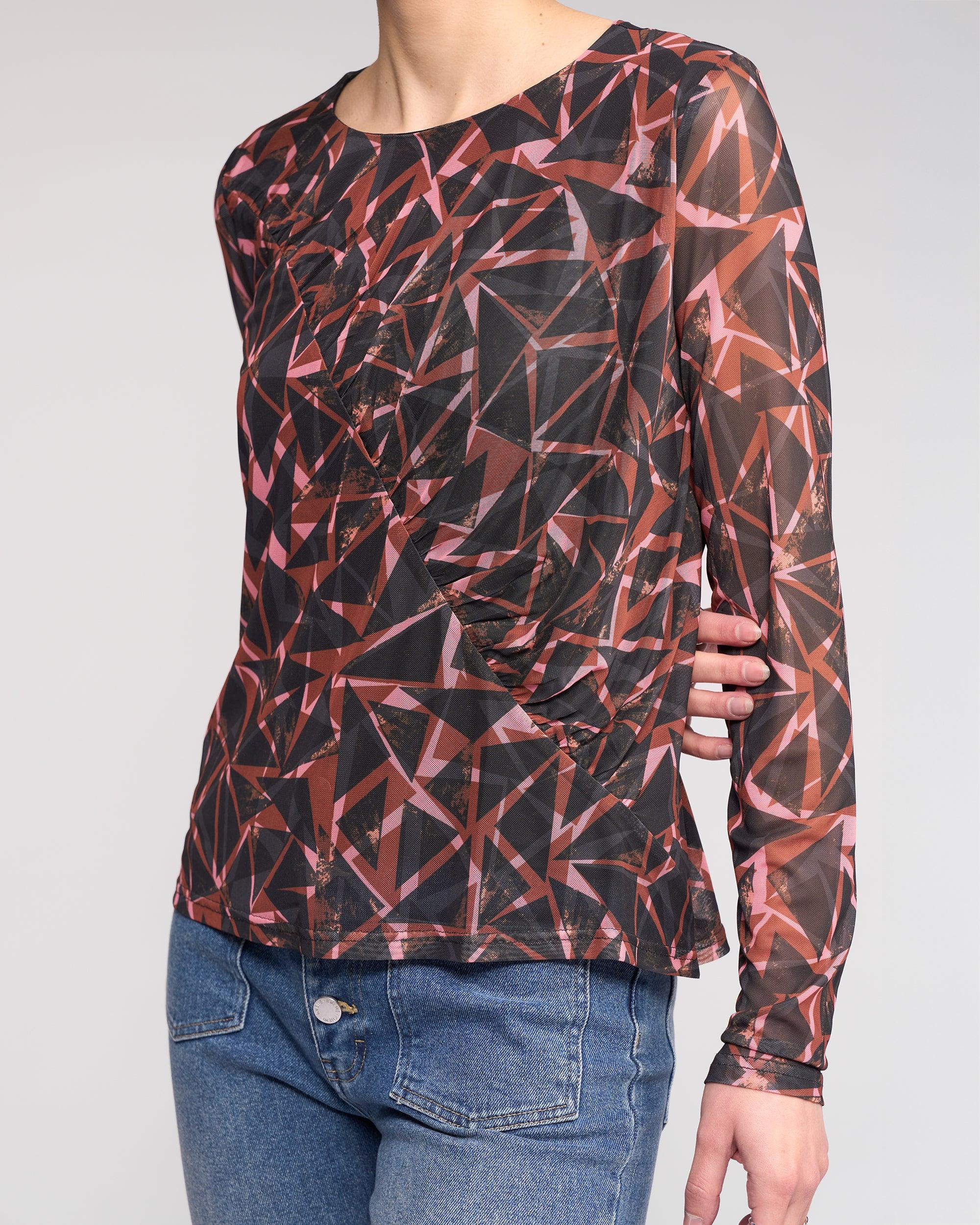 A person wears the Knewe Label Apex Top - Dusk, a long sleeve mesh top in a geometric red, pink, and black print, paired with blue jeans. Their head is out of frame and one hand rests on their hip.