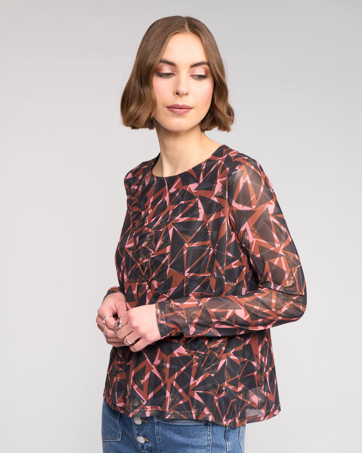 A woman with short brown hair wears the Knewe Label Apex Top - Dusk, a long sleeve mesh top with a pink and black geometric print, as she stands against a light gray background looking slightly to the side.