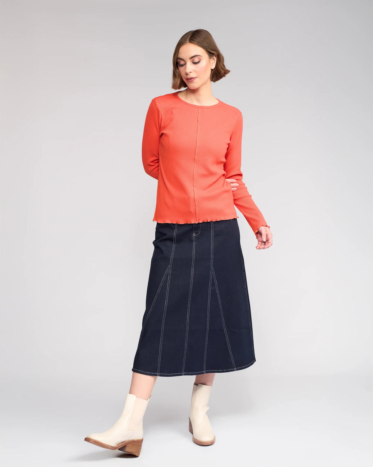 A woman stands against a plain background wearing the Knewe Label Sofie Top in Lava, a dark blue midi denim skirt with visible seams, and light ankle boots. She looks slightly downward with one foot forward.