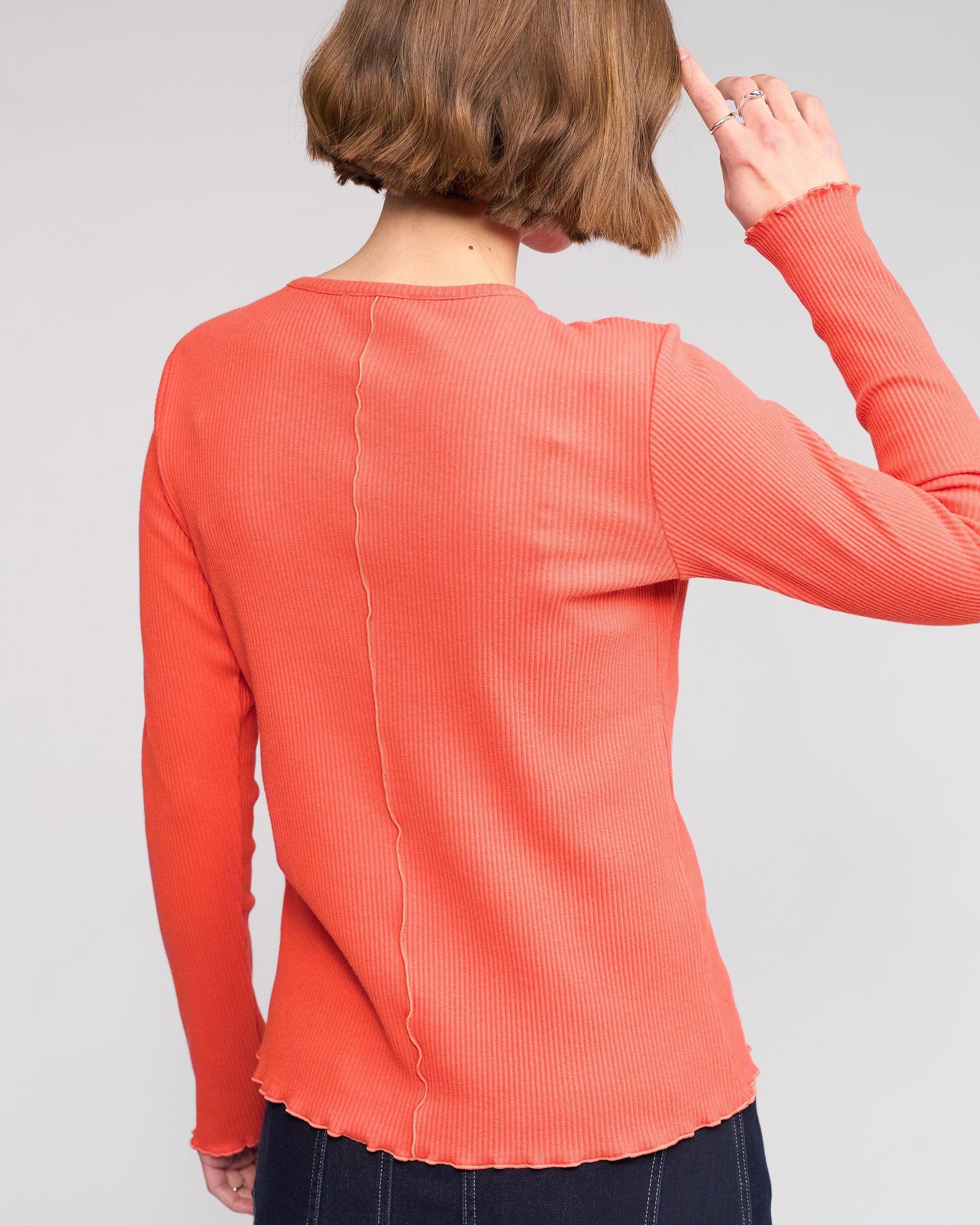 Seen from behind, a person with short brown hair wears the Knewe Label Sofie Top - Lava, a long sleeve orange ribbed cotton top with visible seam details and lettuce-edge seams down the center back and sleeves. One hand is near their head.