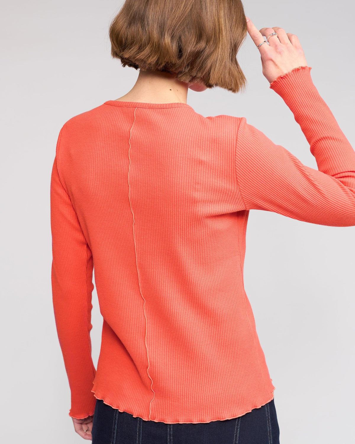 Seen from behind, a person with short brown hair wears the Knewe Label Sofie Top - Lava, a long sleeve orange ribbed cotton top with visible seam details and lettuce-edge seams down the center back and sleeves. One hand is near their head.