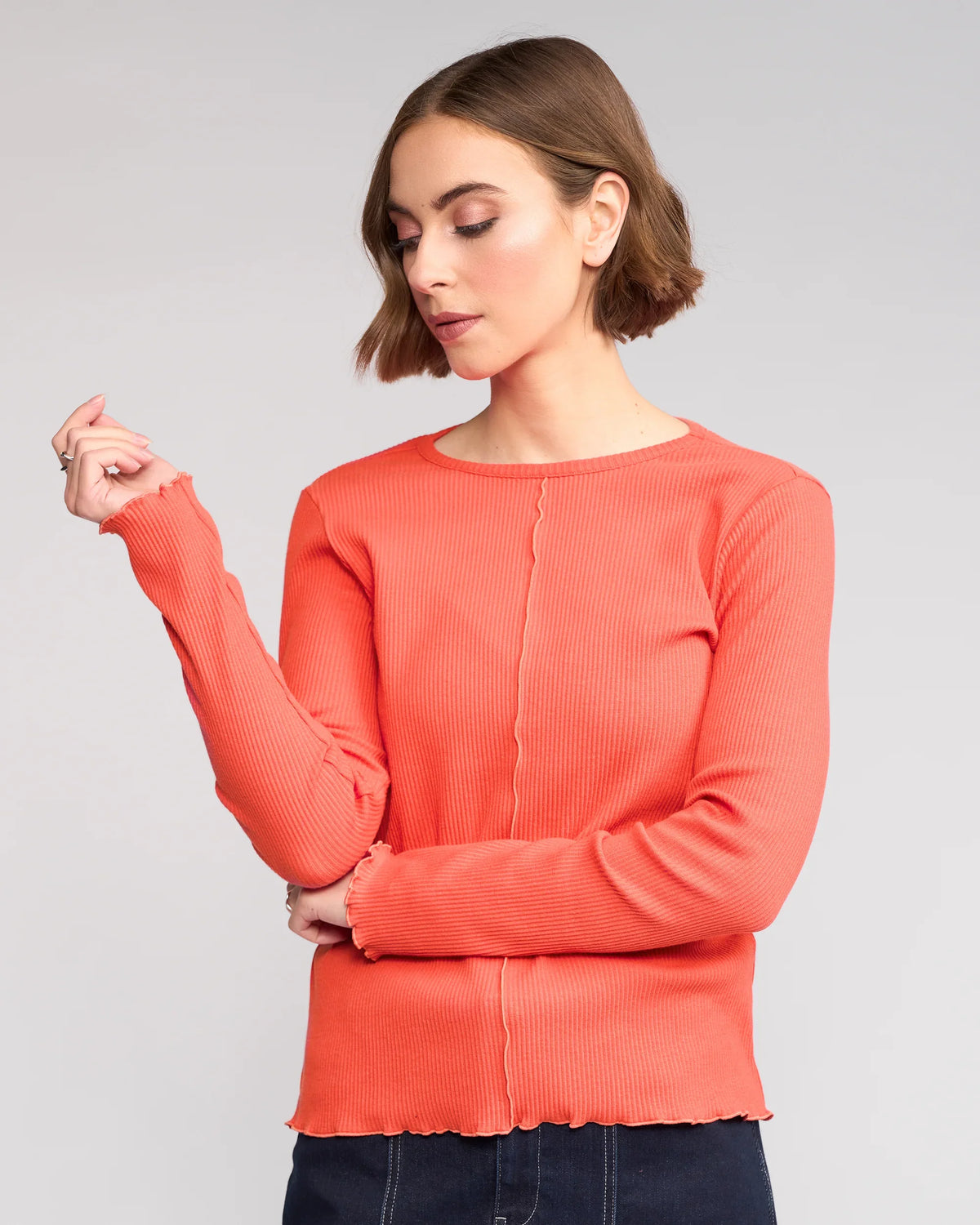 A woman with short brown hair, arms crossed, looks down and to the side against a light gray background. She wears the Knewe Label Sofie Top in Lava—an orange long sleeve top with lettuce-edge seams.