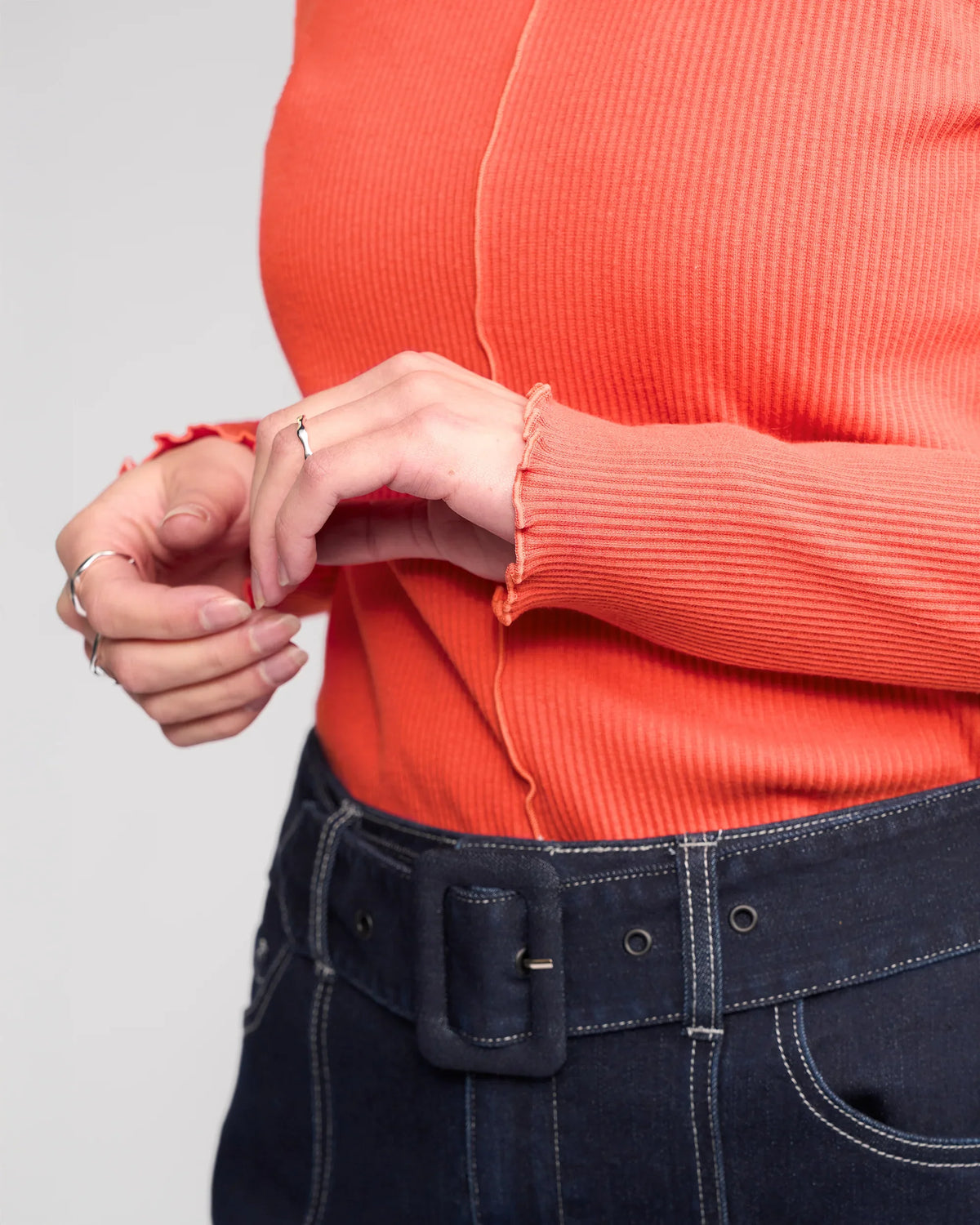A person wearing the Knewe Label Sofie Top in Lava, a long sleeve orange ribbed cotton top with lettuce-edge seams, paired with high-waisted dark denim jeans and silver rings on their hands.