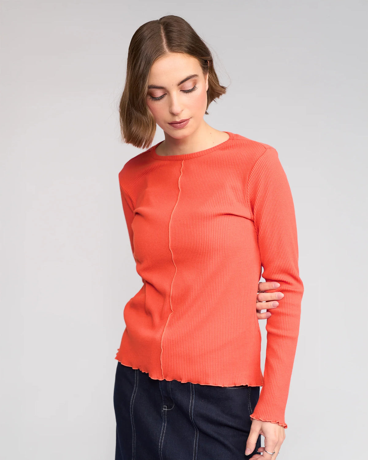 A woman with short brown hair wears the Knewe Label Sofie Top in Lava, featuring long sleeves and lettuce-edge seams, paired with a dark skirt. She stands against a light grey background, looking down with one arm behind her back.