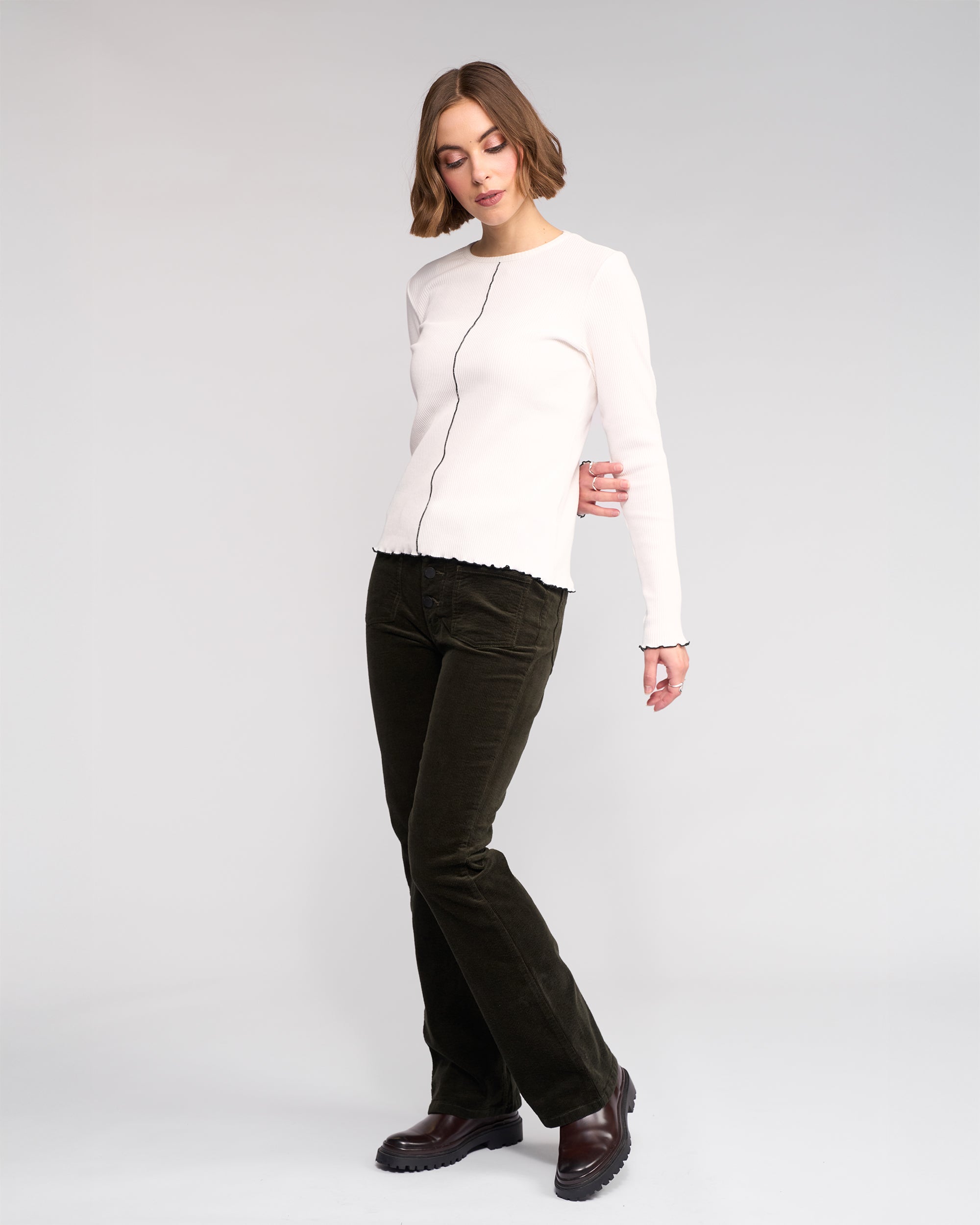 A woman with short, wavy brown hair wears the Knewe Label Sofie Top in Off White, paired with dark green corduroy pants and brown shoes, standing against a plain light background.