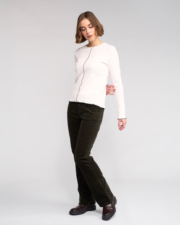 A woman with short, wavy brown hair wears the Knewe Label Sofie Top in Off White, paired with dark green corduroy pants and brown shoes, standing against a plain light background.