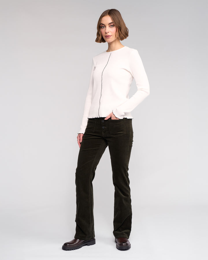 A woman with short hair and a neutral expression stands against a plain background, wearing the Knewe Label Sofie Top in Off White, paired with dark corduroy pants and black shoes. One hand is in her pocket.