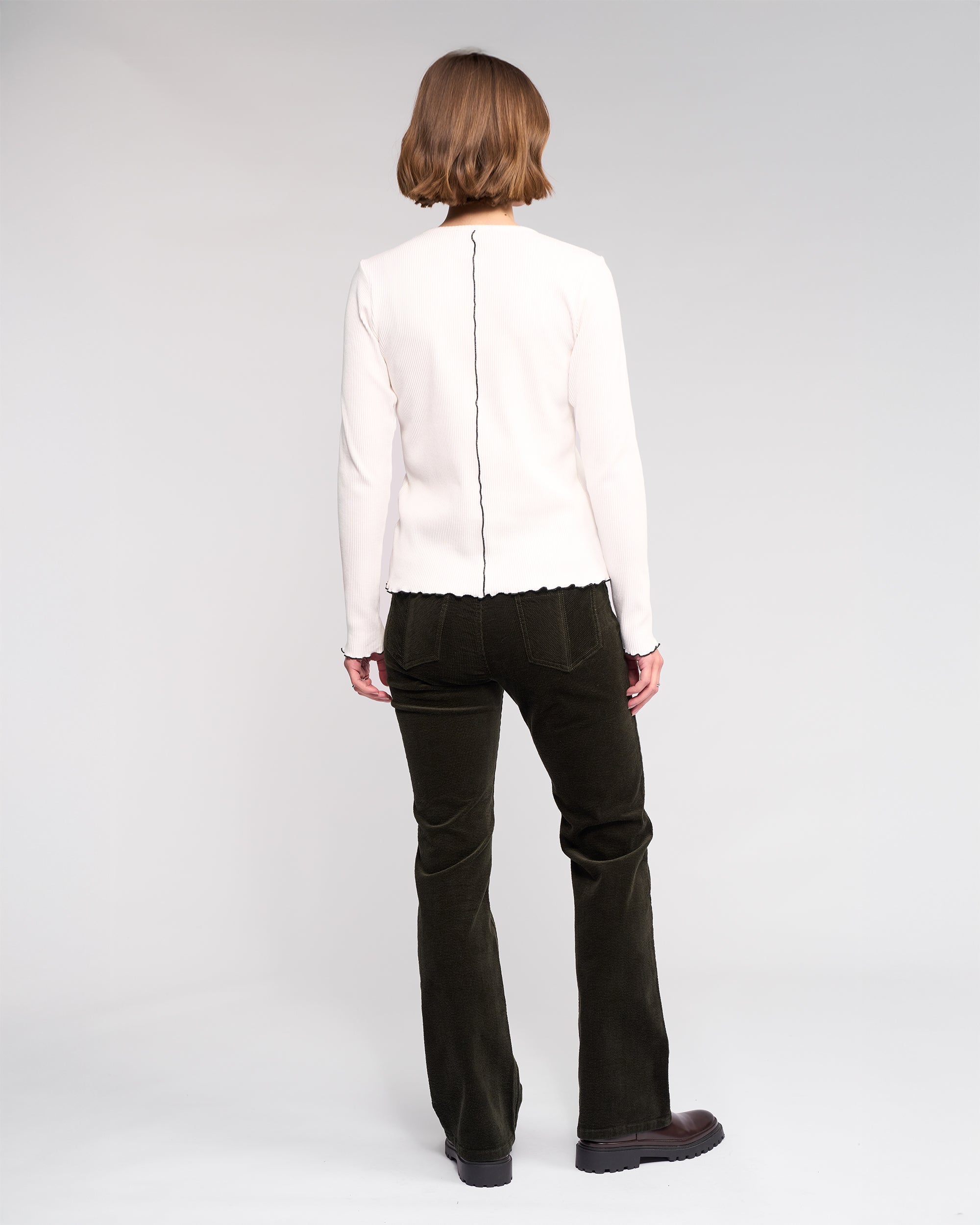 A person with short hair, seen from behind, wears the Knewe Label Sofie Top in Off White—a long sleeve ribbed cotton top with a central seam—paired with black pants against a plain light background.