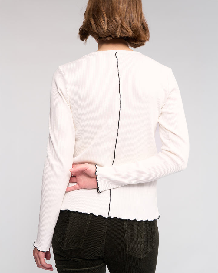 A person with short brown hair, seen from behind, wears the Sofie Top in Off White by Knewe Label—featuring long sleeves, black trim, and ribbed cotton with lettuce-edge seams—paired with dark pants; one arm is bent behind their back.