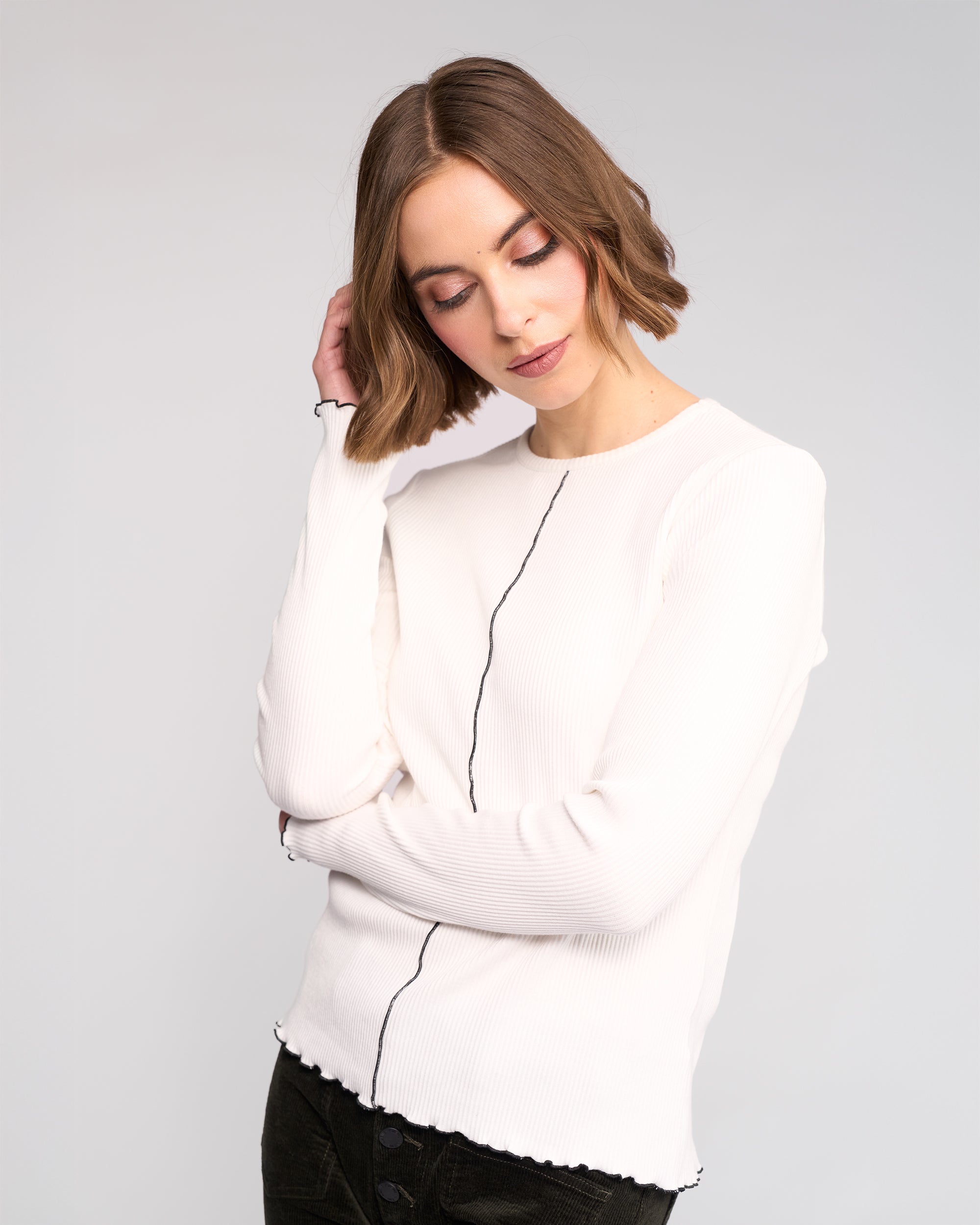 A woman with a bob haircut wears the Knewe Label Sofie Top in off white and dark pants. She stands against a plain background, tilting her head and touching her hair while looking down.