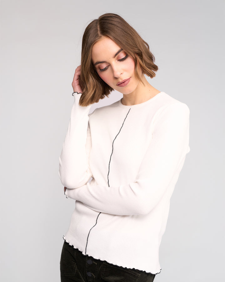 A woman with a bob haircut wears the Knewe Label Sofie Top in off white and dark pants. She stands against a plain background, tilting her head and touching her hair while looking down.