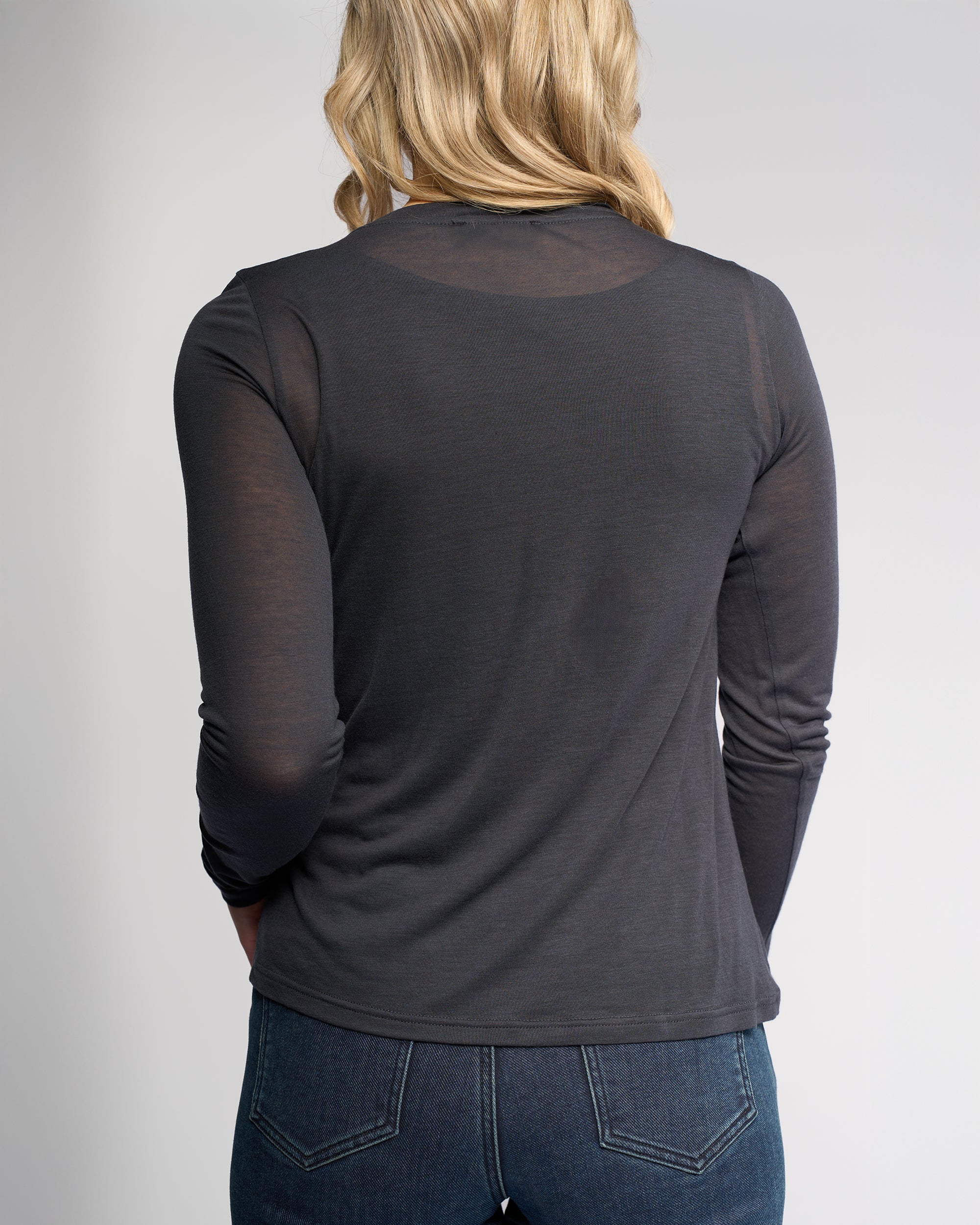 A person with blonde hair is shown from the back, wearing Knewe Label’s Hero Top in Airforce—a blue-grey, long sleeve Tencel wool blend—paired with dark blue jeans against a plain light background.