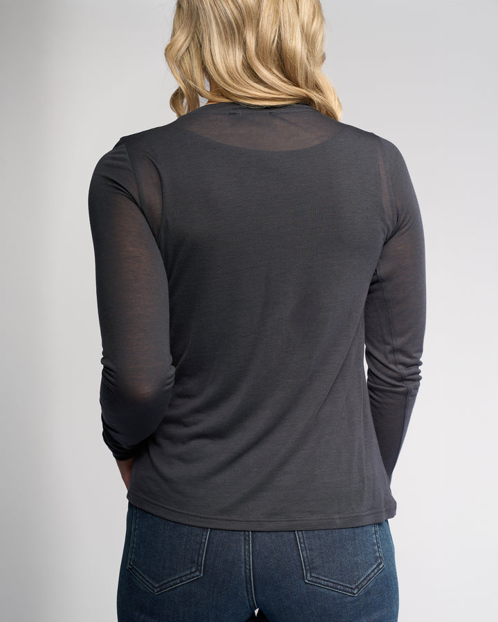 A person with blonde hair is shown from the back, wearing Knewe Label’s Hero Top in Airforce—a blue-grey, long sleeve Tencel wool blend—paired with dark blue jeans against a plain light background.