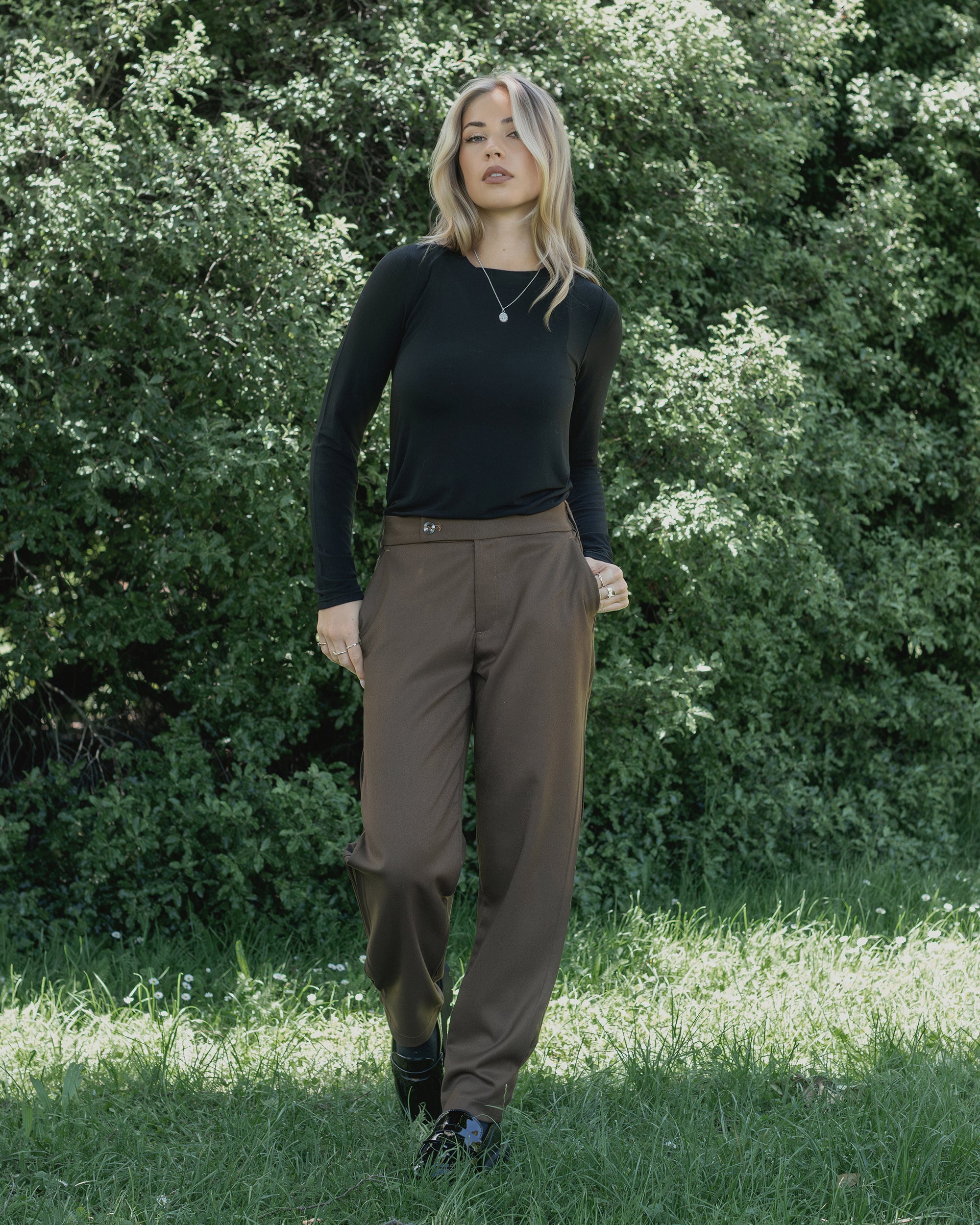 A woman with wavy blonde hair wears the Knewe Label Jude Pant in Cedar and a long-sleeve black top, standing outdoors on grass with green foliage behind her.