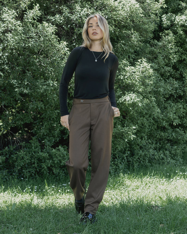 A woman with wavy blonde hair wears the Knewe Label Jude Pant in Cedar and a long-sleeve black top, standing outdoors on grass with green foliage behind her.