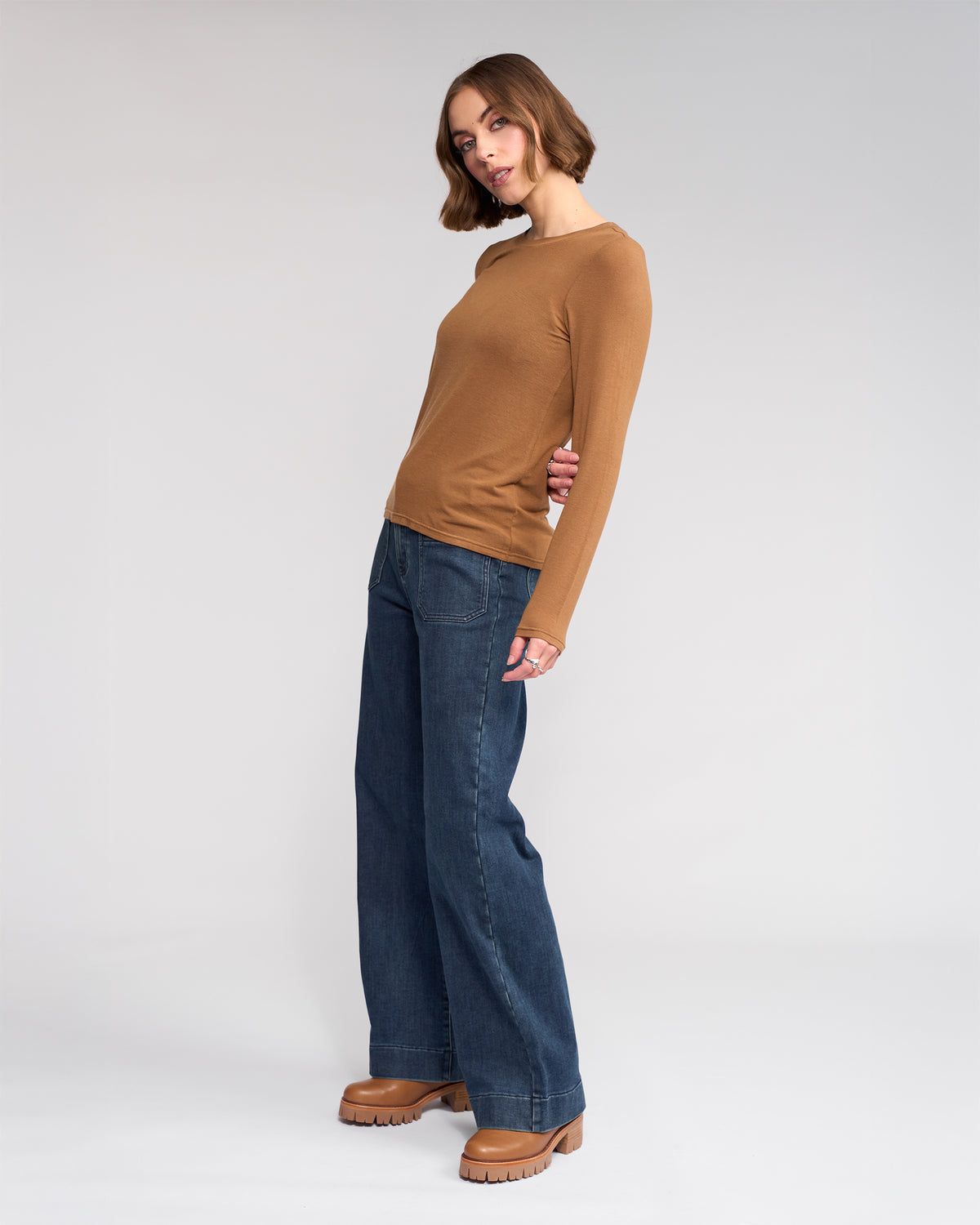 A woman poses against a plain background in the Knewe Label Hero Top - Cedar, a fitted brown long sleeve, paired with wide-leg dark blue jeans and tan platform shoes. She stands slightly angled, one hand at her side, with a relaxed expression.