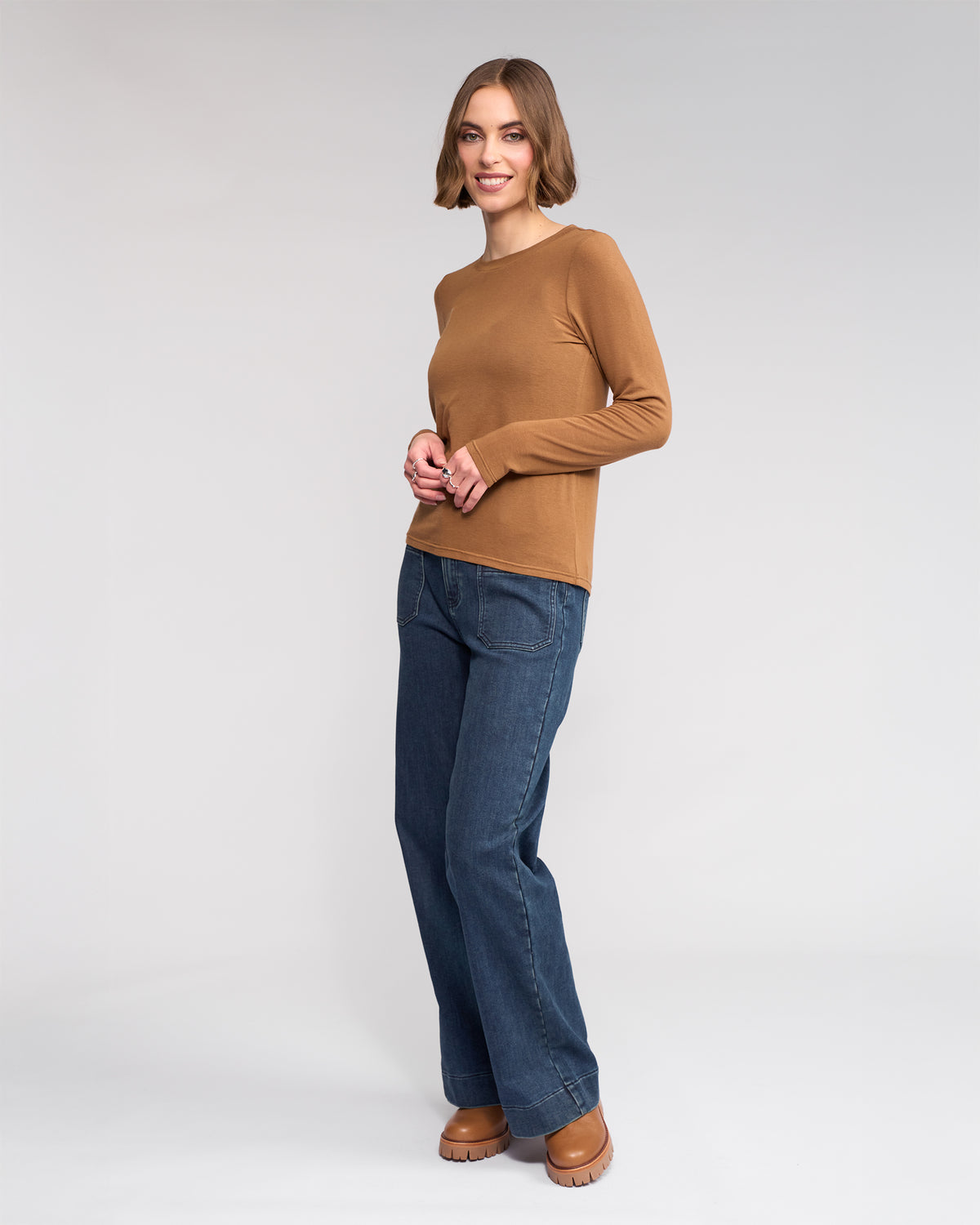 A woman with a bob haircut smiles while standing against a plain light background, wearing the Knewe Label Hero Top in Cedar, blue jeans, and brown platform shoes.