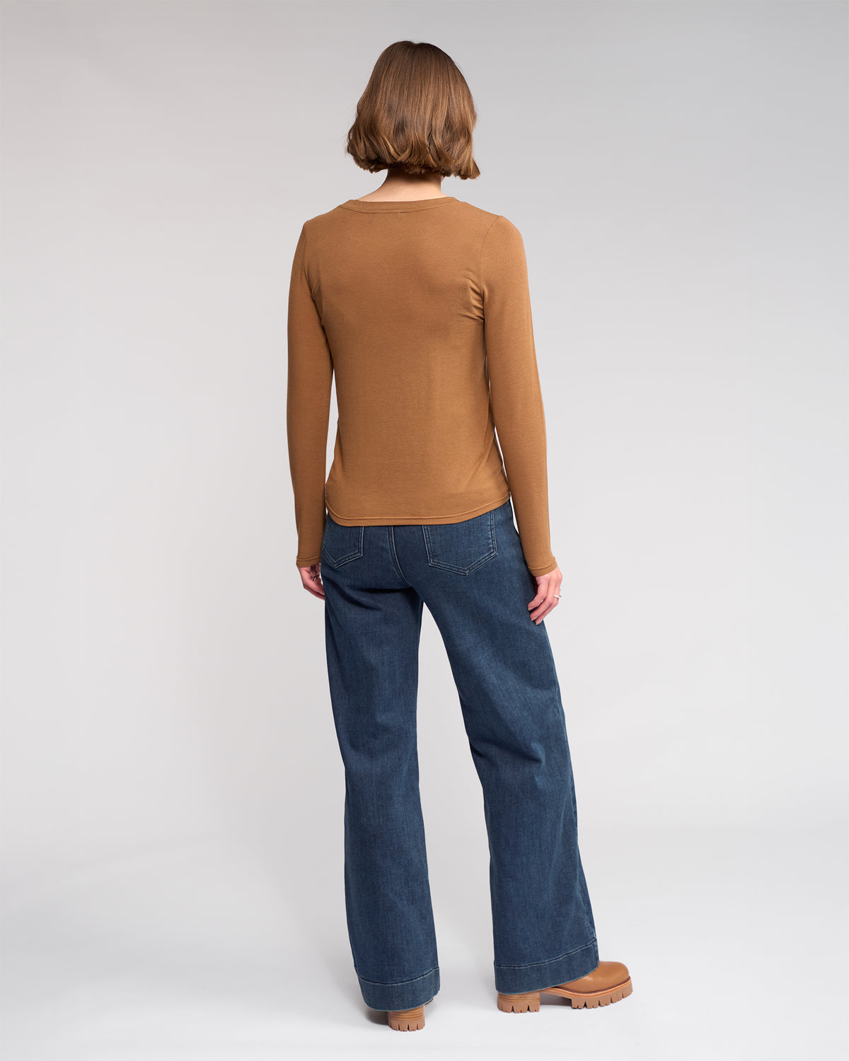 A person with short hair, wearing the Knewe Label Hero Top in Cedar, blue wide-leg jeans, and brown shoes, stands facing away from the camera against a plain light background.