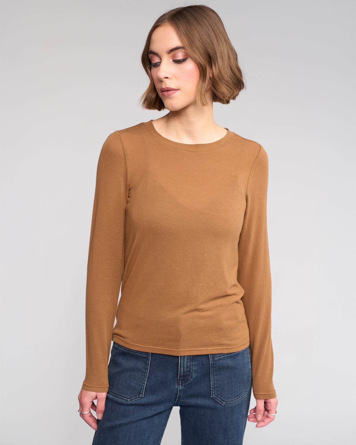 A woman with short brown hair wears the Knewe Label Hero Top in Cedar, a fitted long sleeve layering piece, paired with blue jeans. She stands against a plain light gray background, looking down with a neutral expression.