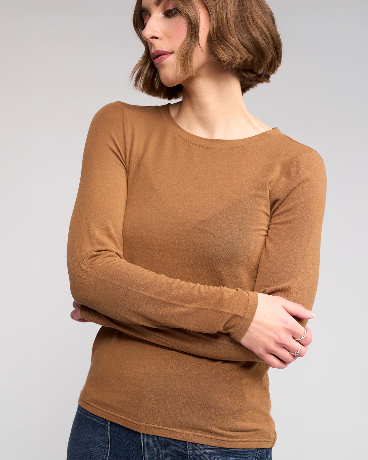 A woman with short brown hair wears the Hero Top in Cedar by Knewe Label and blue jeans, standing with arms crossed and looking to the side against a plain light background.