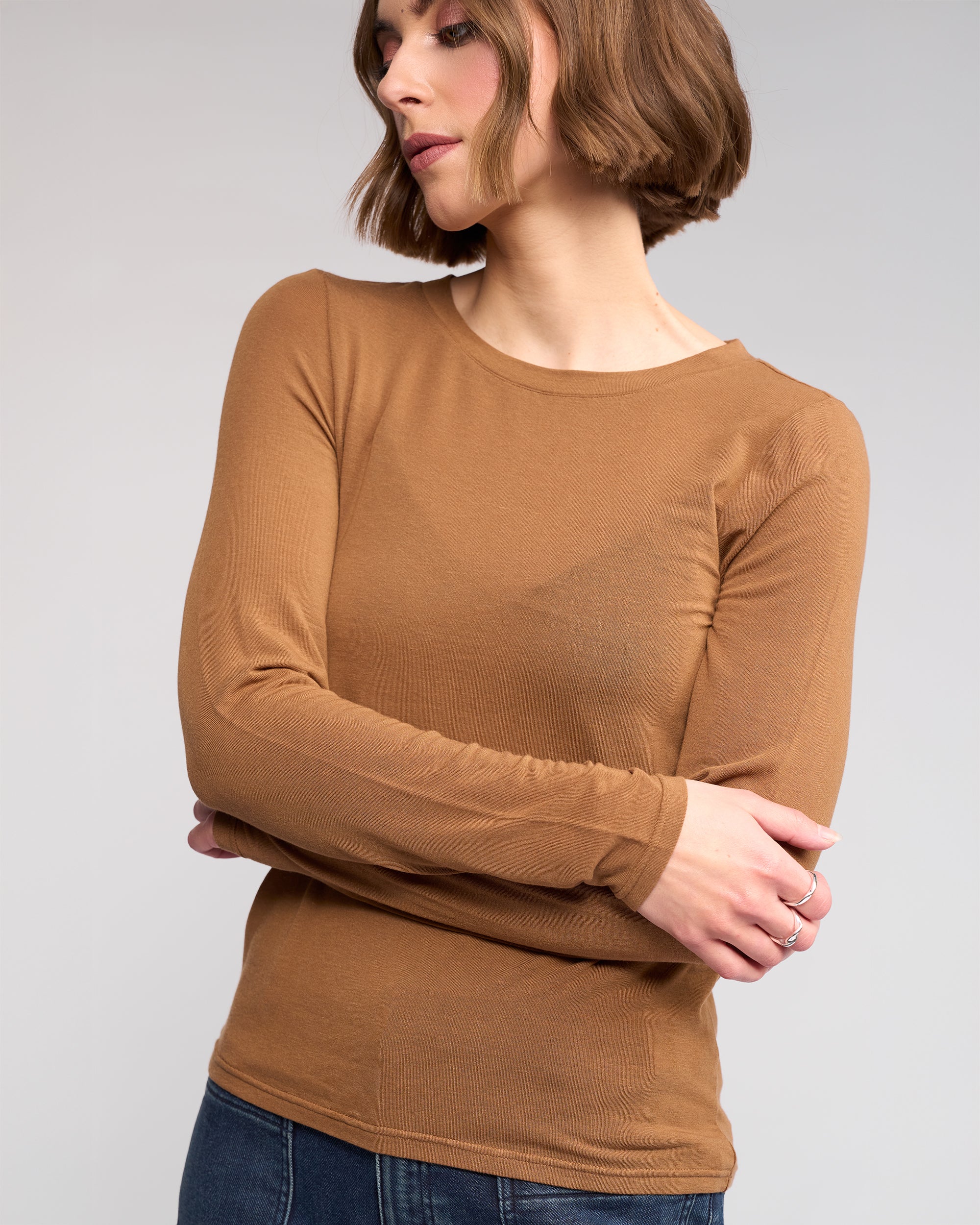 A woman with short brown hair wears the Hero Top in Cedar by Knewe Label and blue jeans, standing with arms crossed and looking to the side against a plain light background.