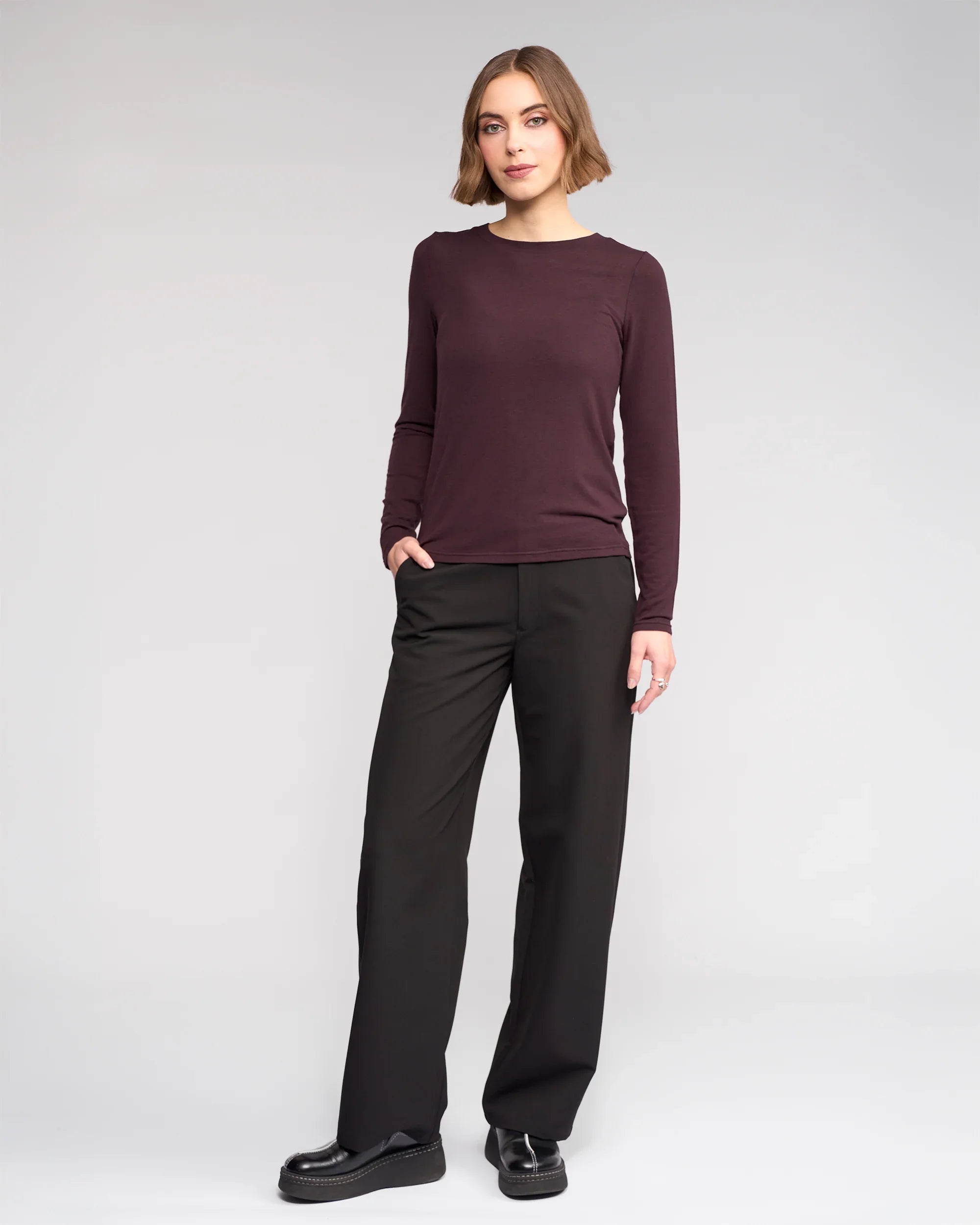 A woman stands against a plain light background, wearing the Knewe Label Hero Top - Raisin, paired with black wide-leg pants and black shoes. One hand is in her pocket as she looks at the camera with a slight smile.