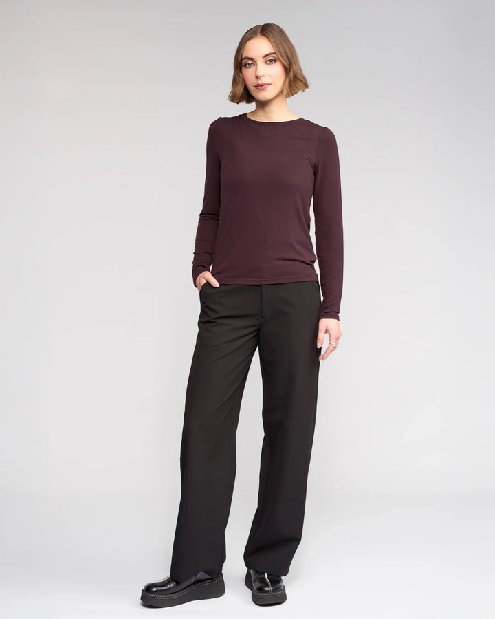 A woman stands against a plain light background, wearing the Knewe Label Hero Top - Raisin, paired with black wide-leg pants and black shoes. One hand is in her pocket as she looks at the camera with a slight smile.