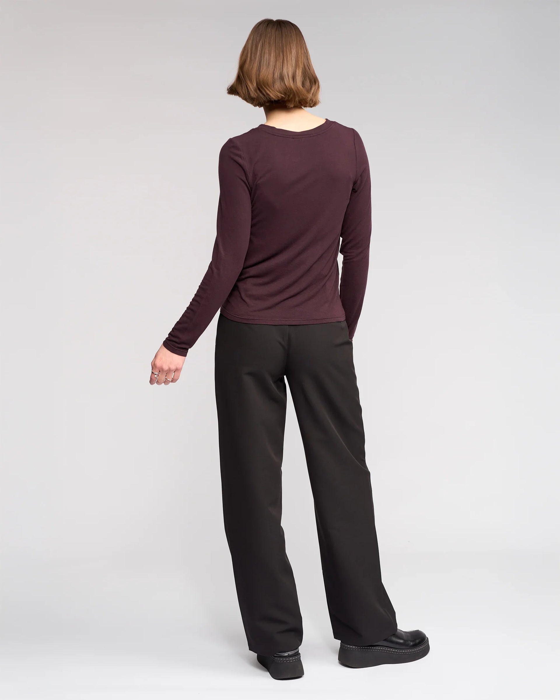 A person with short brown hair faces away, wearing the Knewe Label Hero Top in Raisin, black wide-leg pants, and black platform shoes against a plain light gray background.