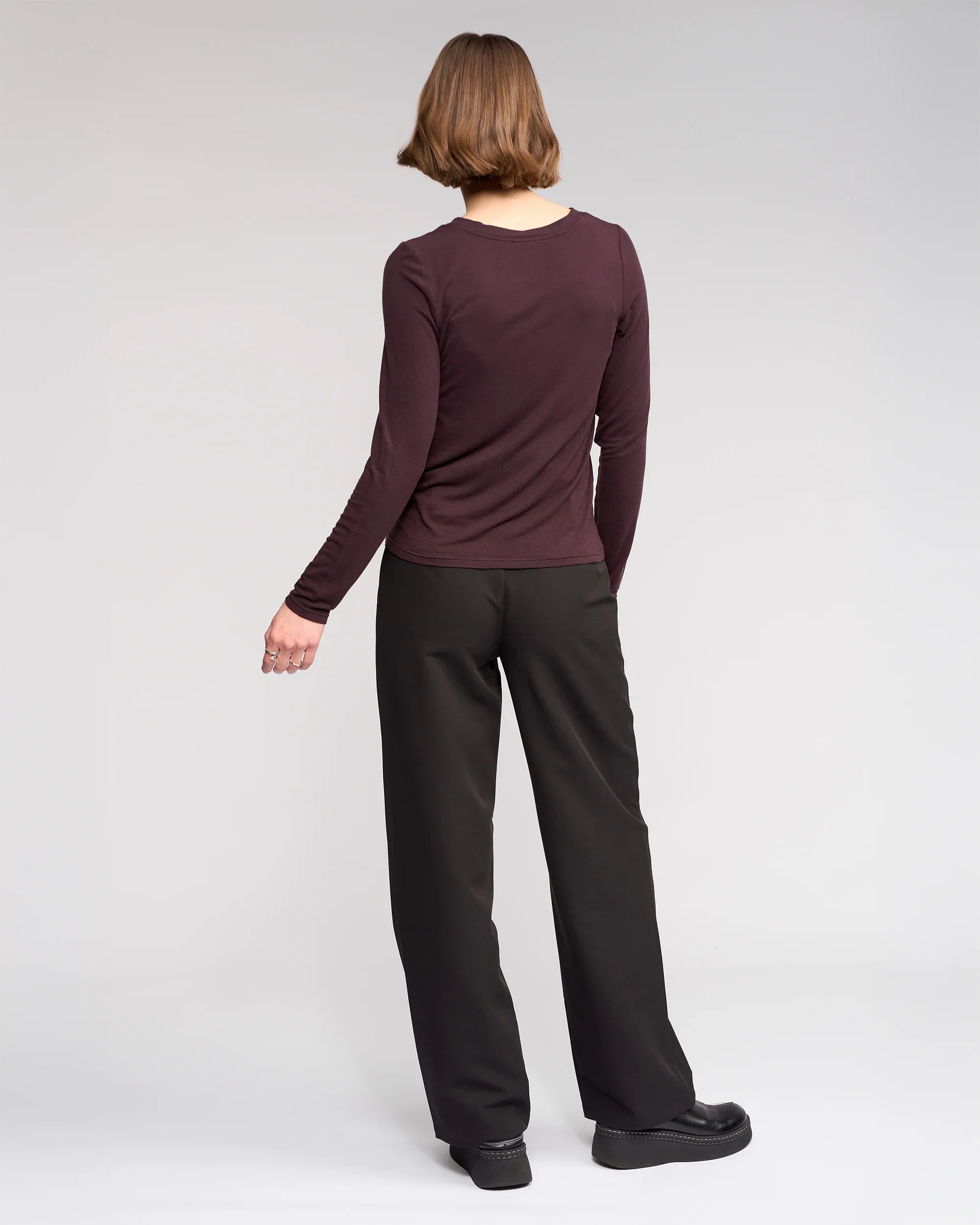 A person with short brown hair faces away, wearing the Knewe Label Hero Top in Raisin, black wide-leg pants, and black platform shoes against a plain light gray background.